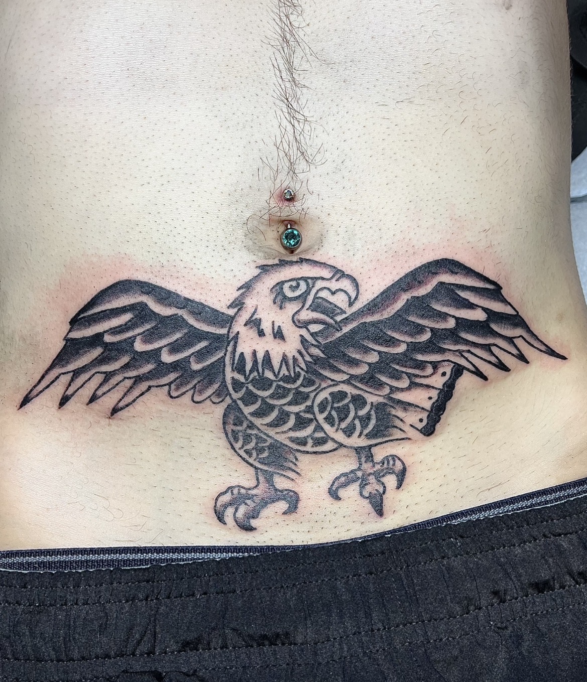 Tattoo of an eagle with outstretched wings on person's lower abdomen, above the waistband, with a piercing above it.