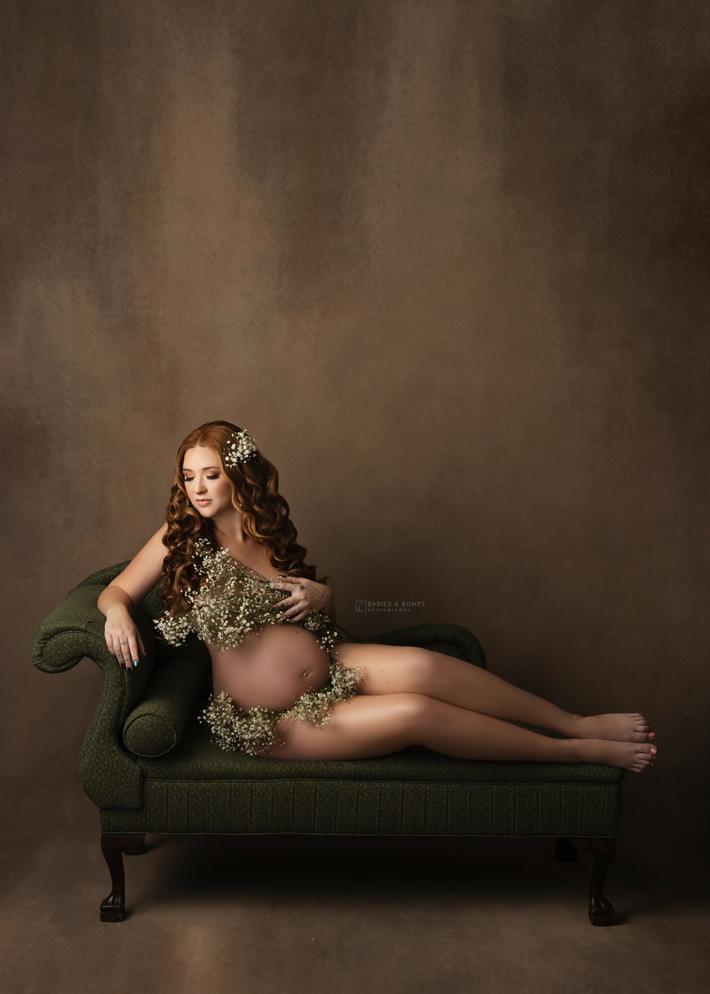Woman with long hair lying on a green sofa, wearing a floral dress, against a plain brown background.