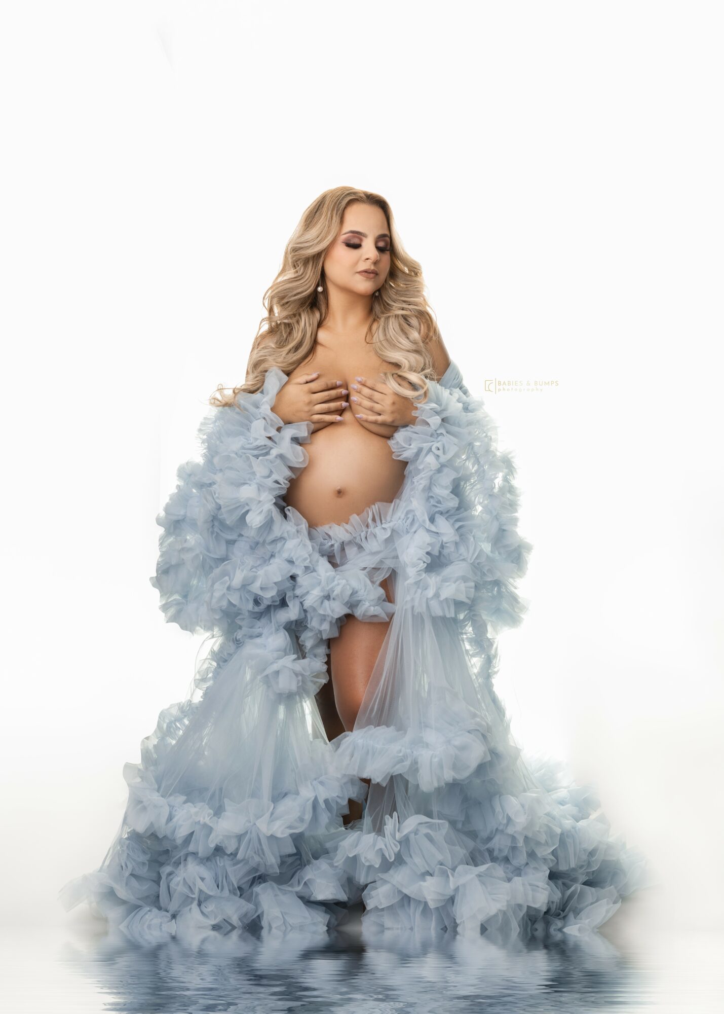 Pregnant woman with long wavy hair wearing a fluffy blue gown, holding her belly, against a white background.