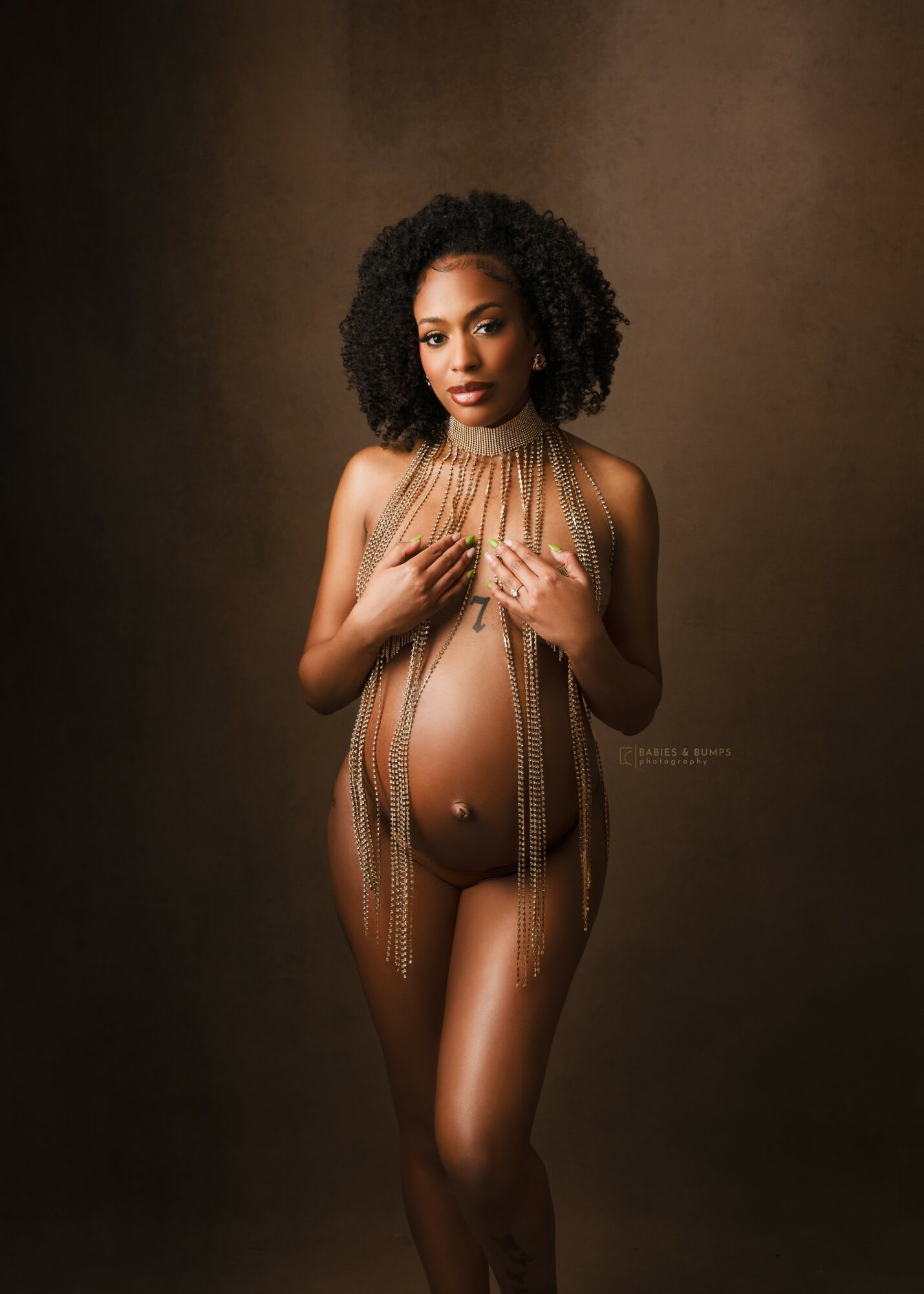 Pregnant woman with curly hair poses against a brown background, covering her chest with hands, wearing minimal clothing.