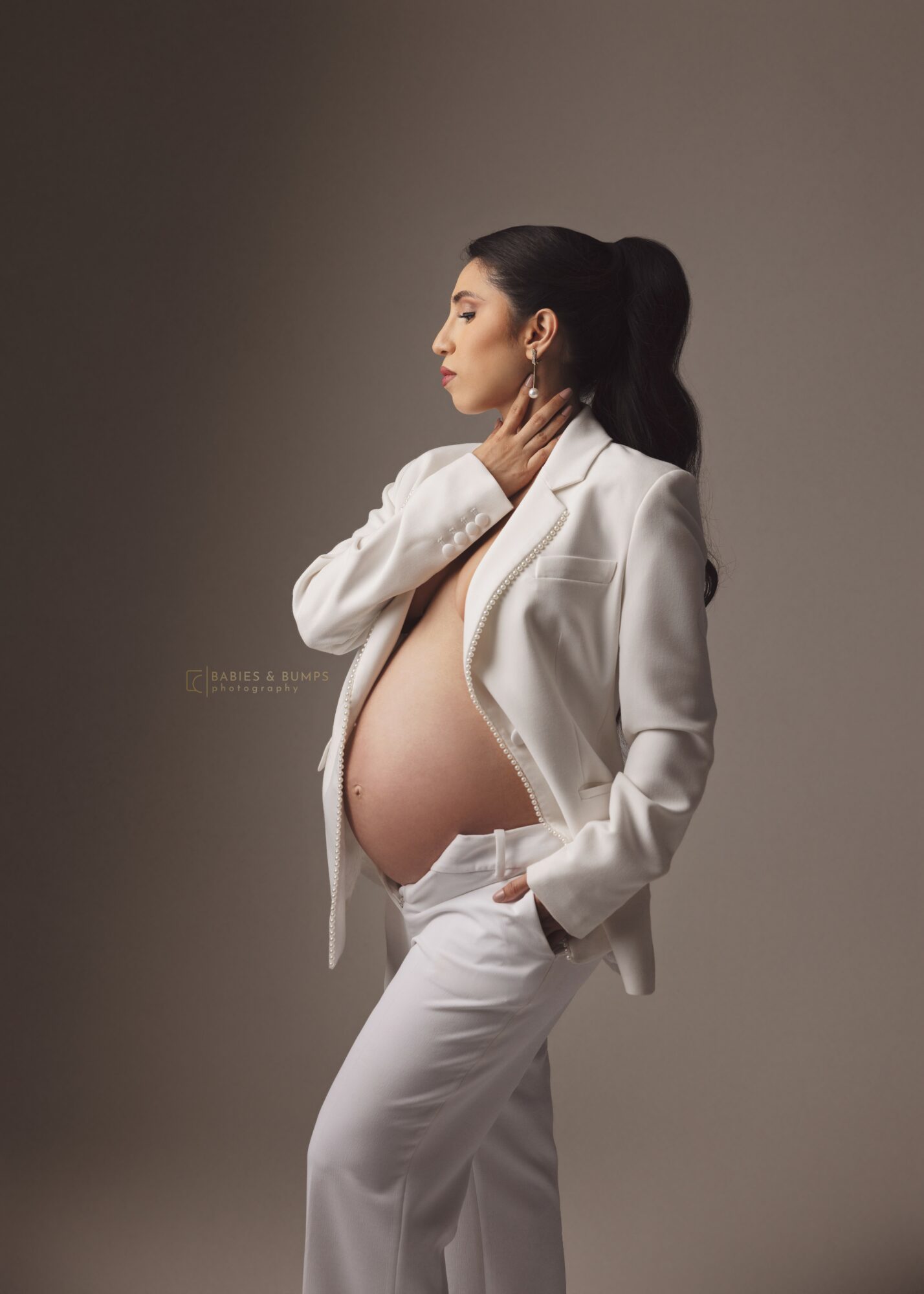 Pregnant woman in white blazer and pants, standing sideways with hand on neck, against neutral background.
