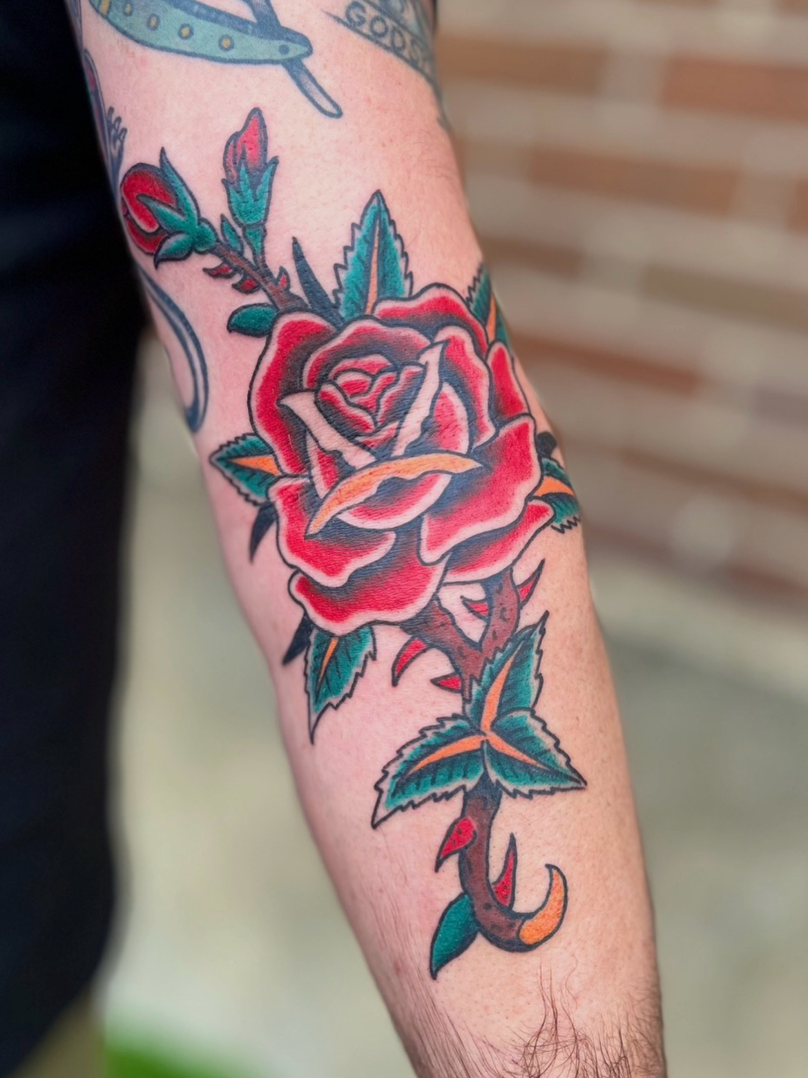 Tattoo of a red rose with green leaves on a person's arm, with a small umbrella hanging below the flower.
