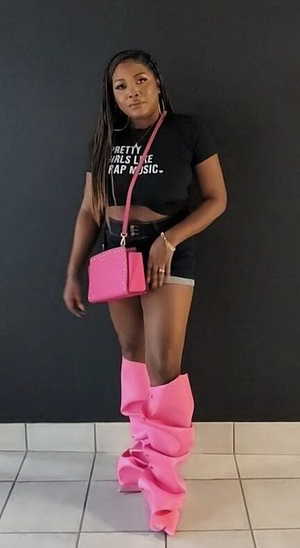 Young woman with long dark hair wearing a black t-shirt, shorts, pink bag, and pink leg coverings, standing against a dark wall.