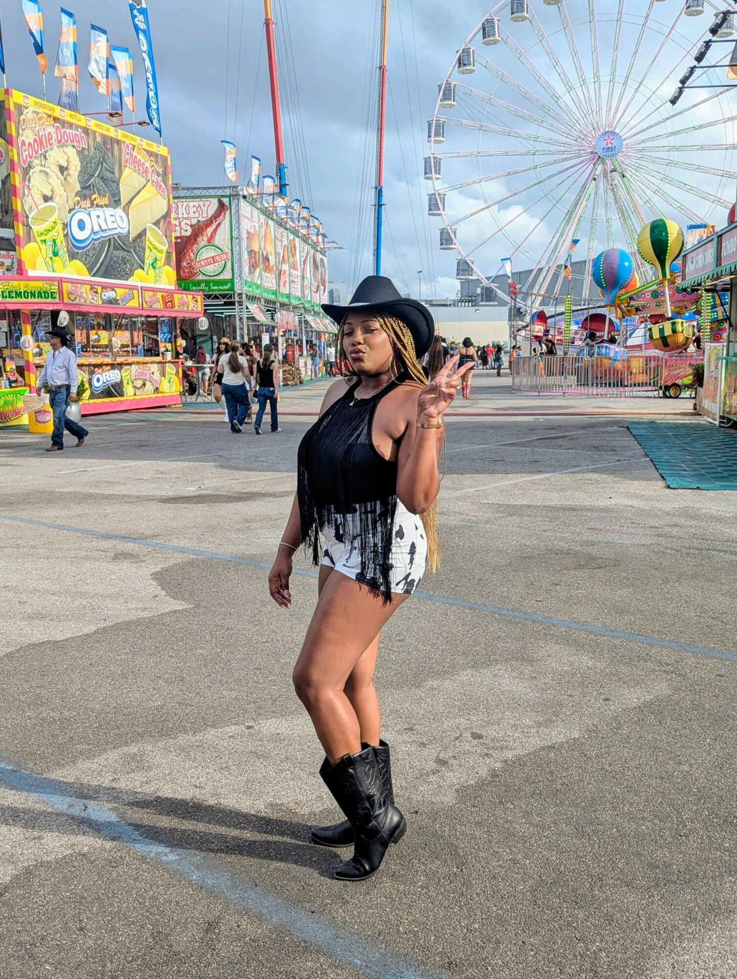 Woman in black hat and boots posing at amusement park with Ferris wheel and rides in background.