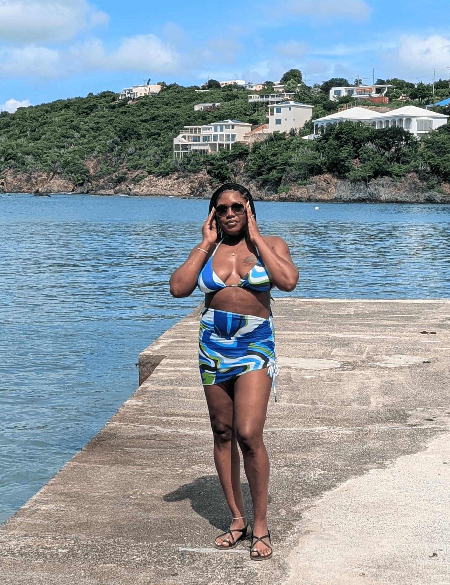 Woman in a blue and white bikini walking on a concrete pier by water, with houses on a hill in the background.