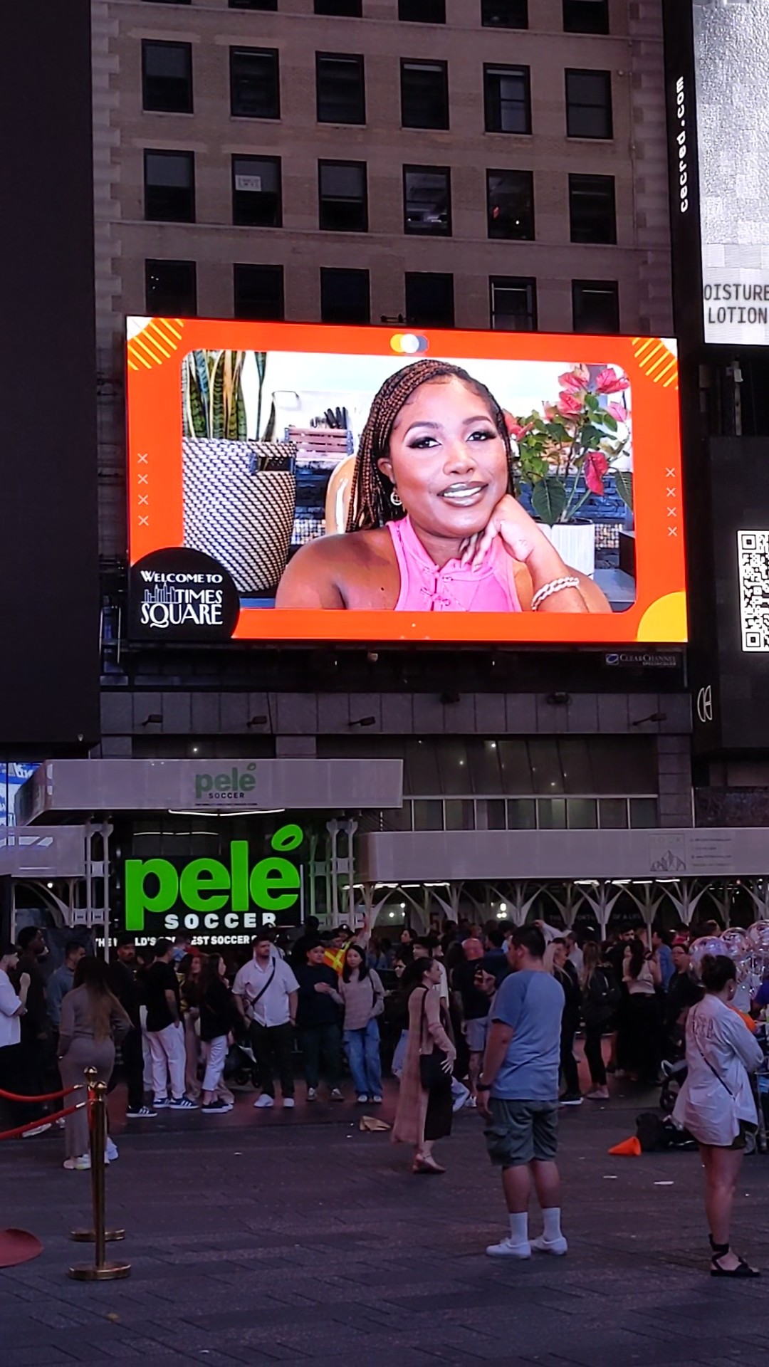 Large digital billboard showing a smiling woman with long hair, surrounded by a red border, above a crowd in an urban area.