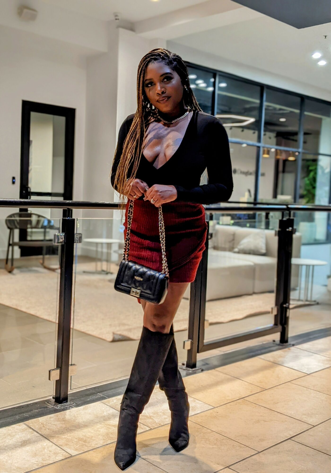 Young woman with long braids in a black top and red skirt, holding a small purse, standing indoors near a glass railing.