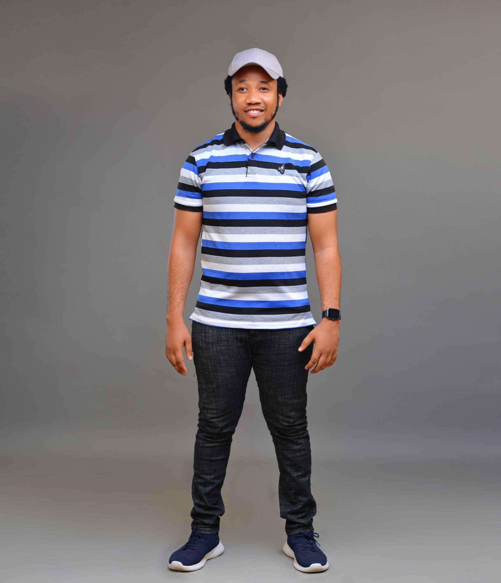 Young man standing against a plain gray background, wearing a striped polo shirt, dark pants, sneakers, and a cap.