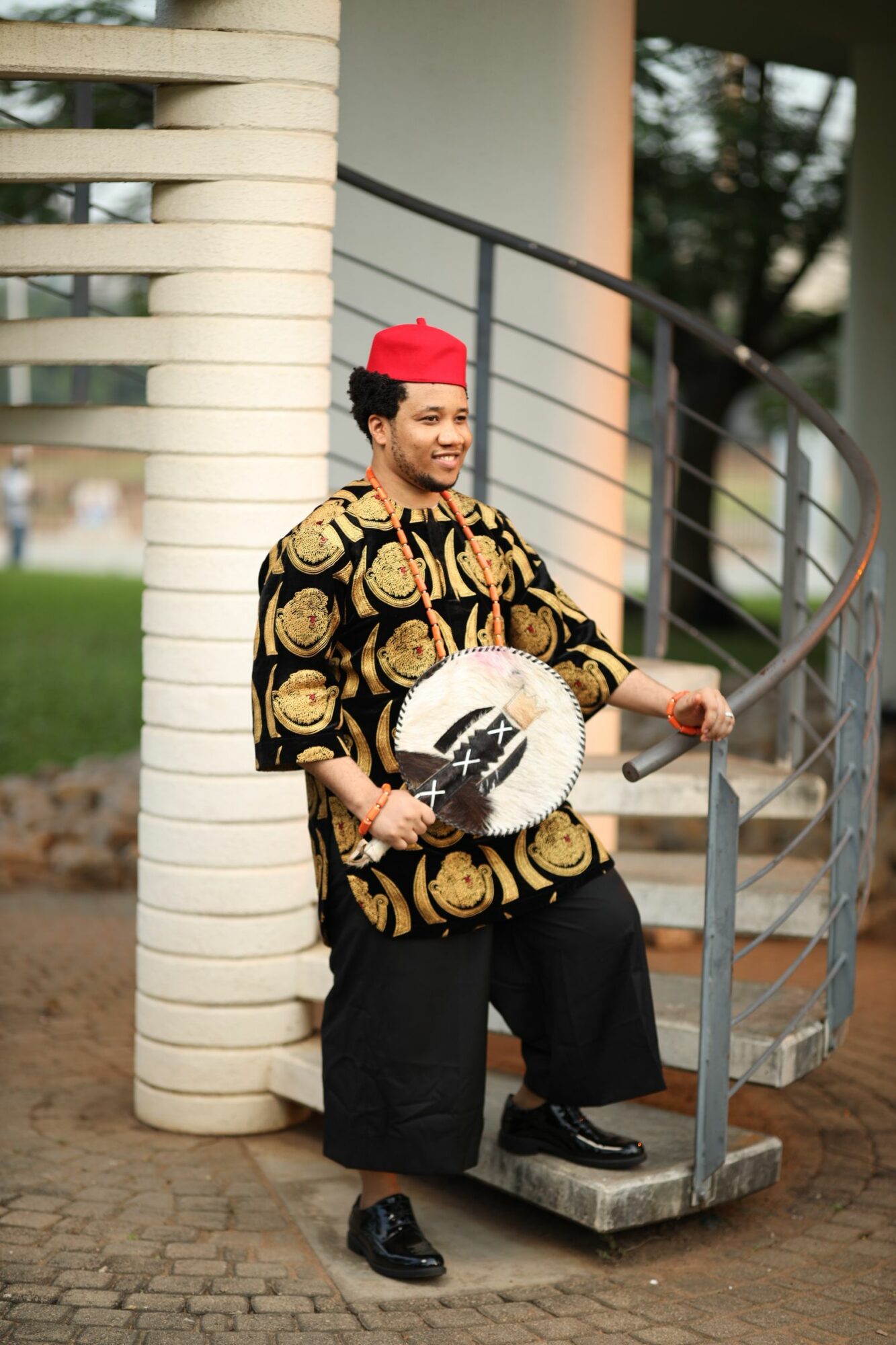 Person dressed in patterned clothing and red hat holding a white object, standing outdoors near a curved metal structure.