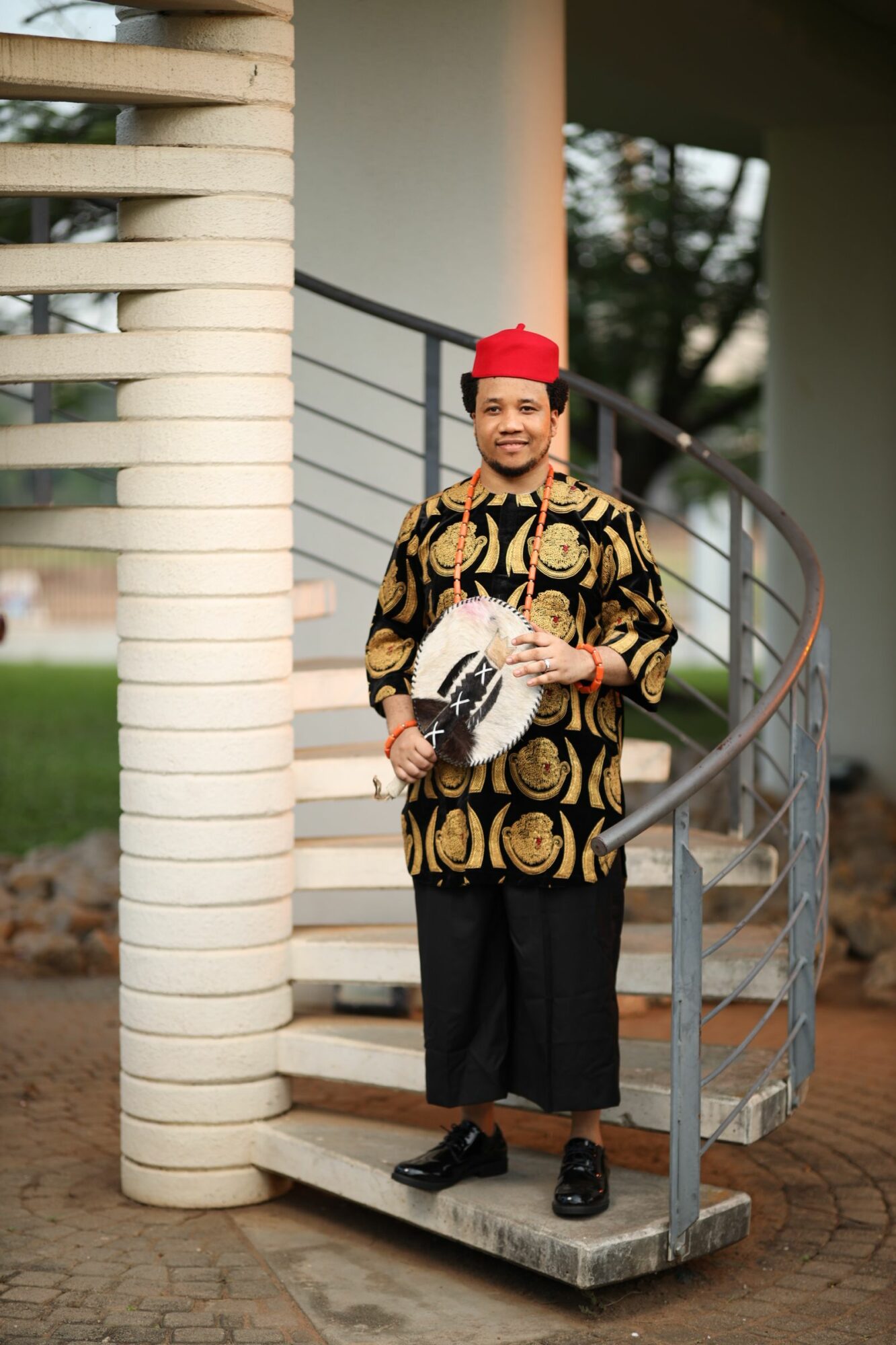 Person sitting on a bench holding a book, wearing a patterned coat and red hat, outdoors with trees and a building.