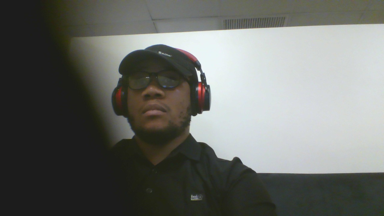Person wearing sunglasses, headphones, and a cap, sitting against a plain wall.