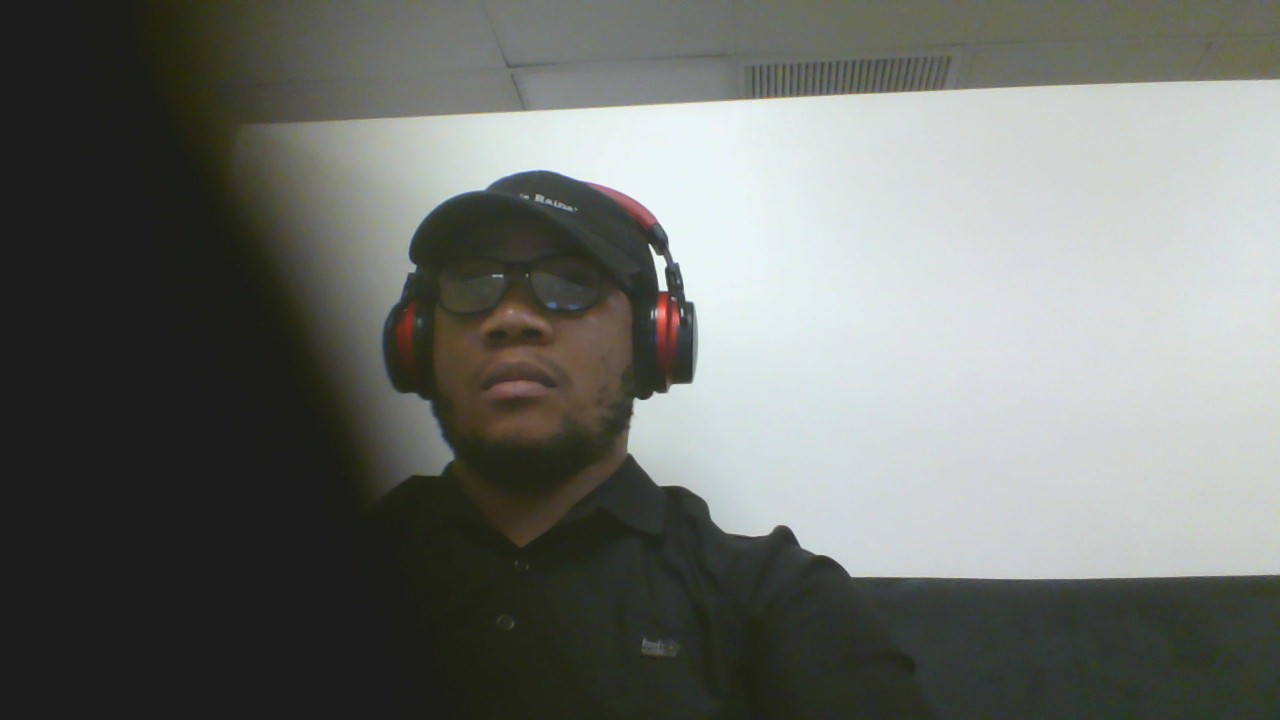 Person wearing headphones and sunglasses, dressed in dark clothing, sitting against a white wall.