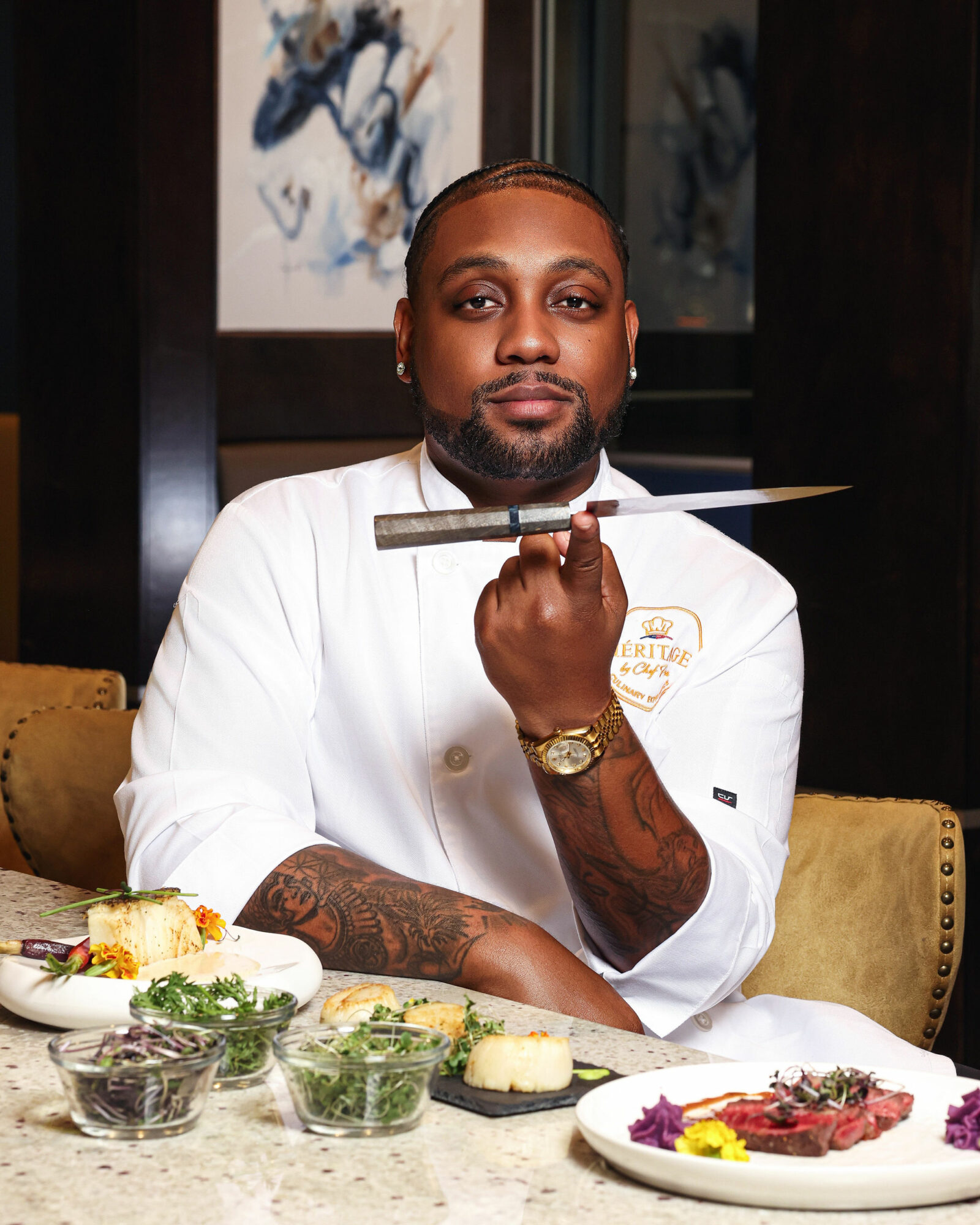 Chef in white uniform holding a knife, seated at a table with food, in a restaurant setting.