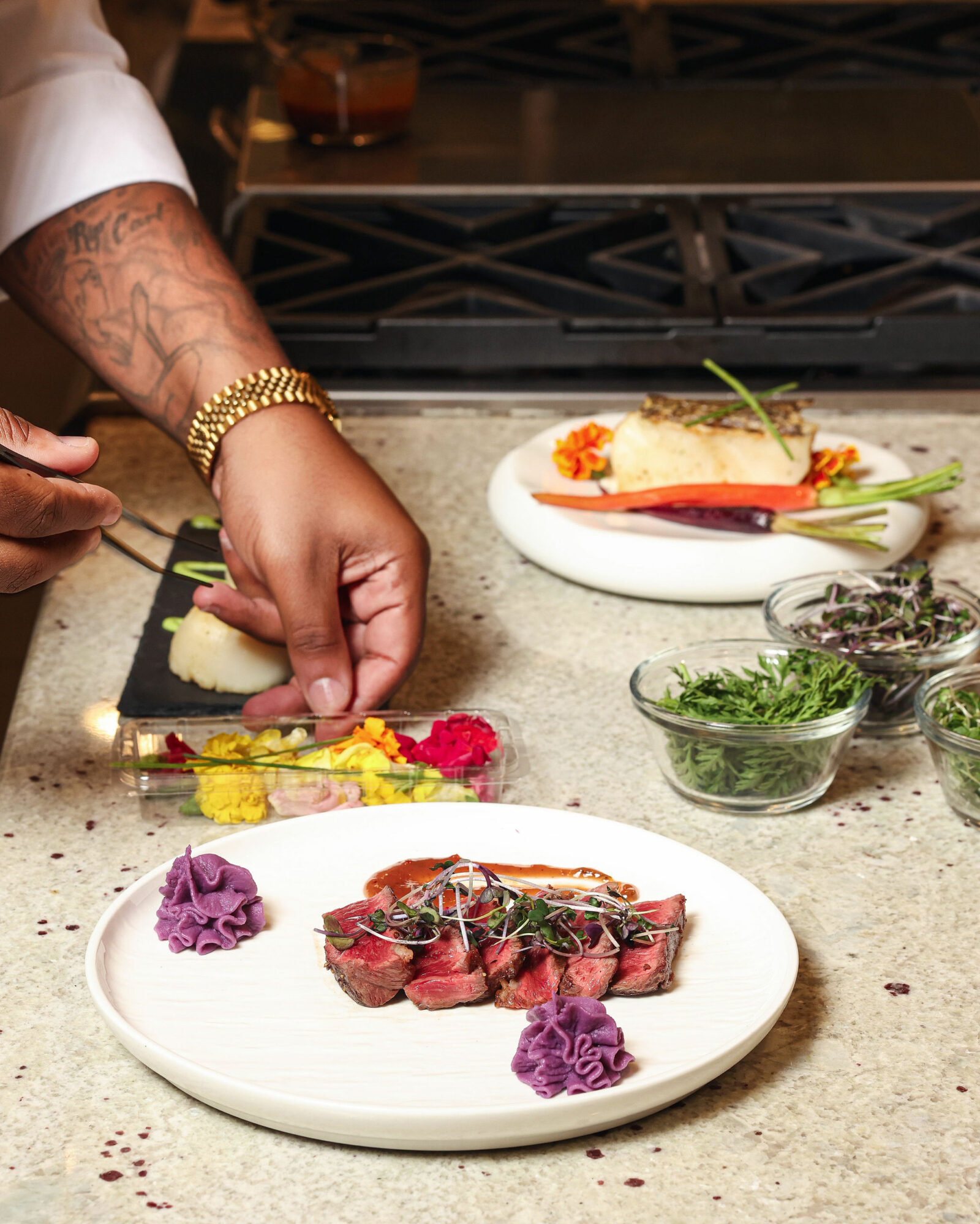 Person preparing a dish with sliced meat, garnished with purple flowers, on a white plate, with herbs and vegetables nearby.