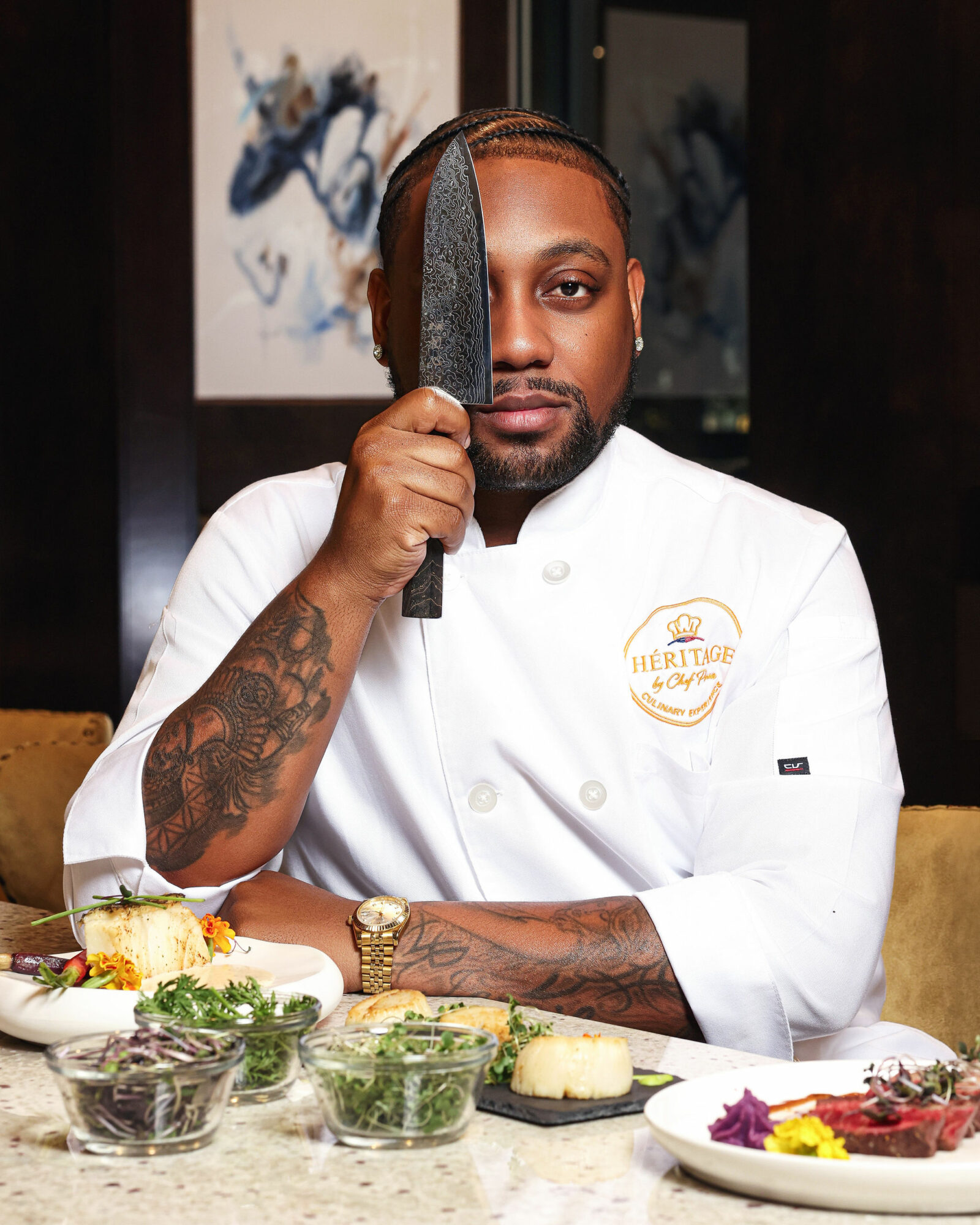 Chef holding a knife vertically in front of face, wearing a white chef's coat with a logo, seated at a table with food.