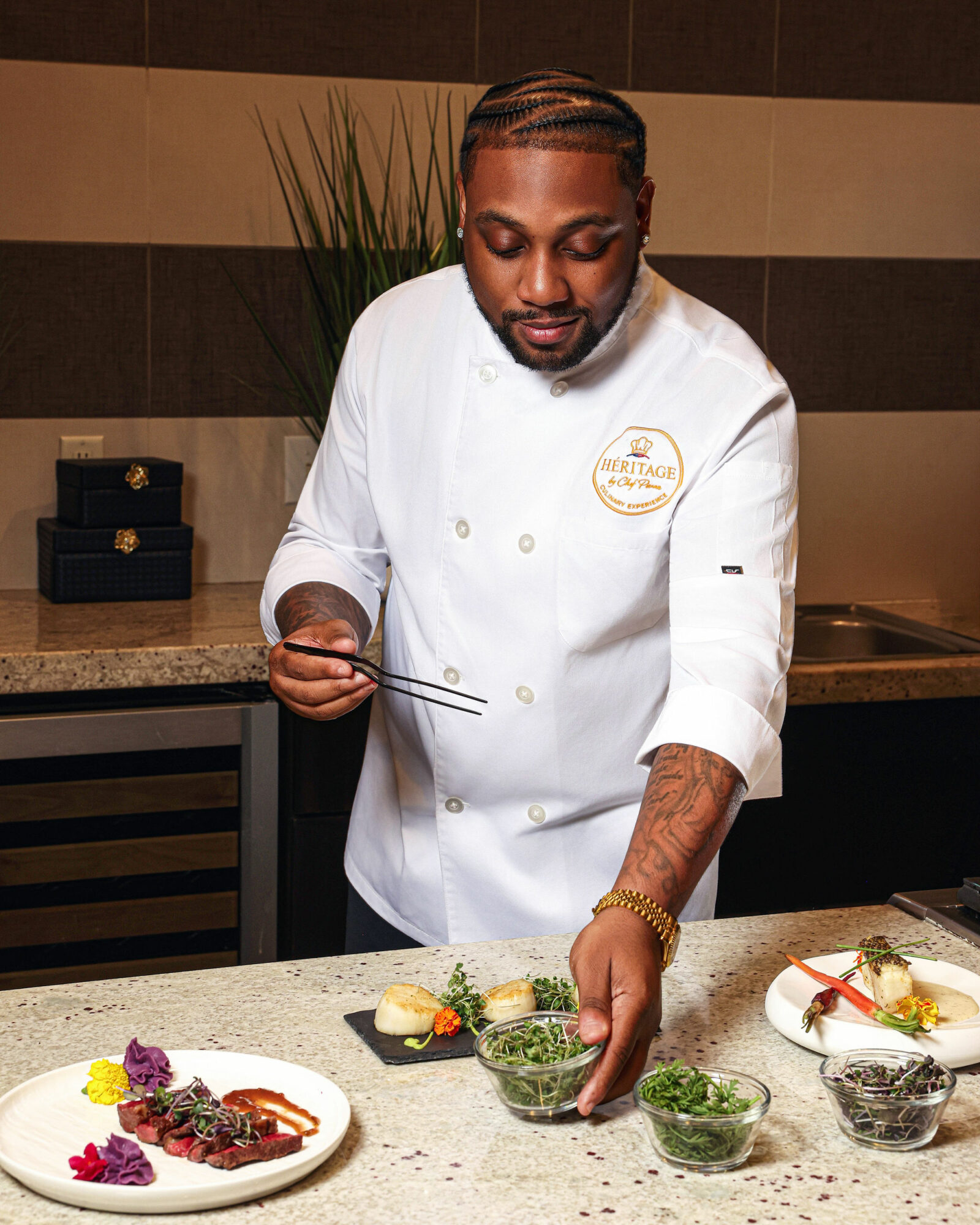 Chef in white uniform preparing dishes with bowls of green herbs on a kitchen counter.