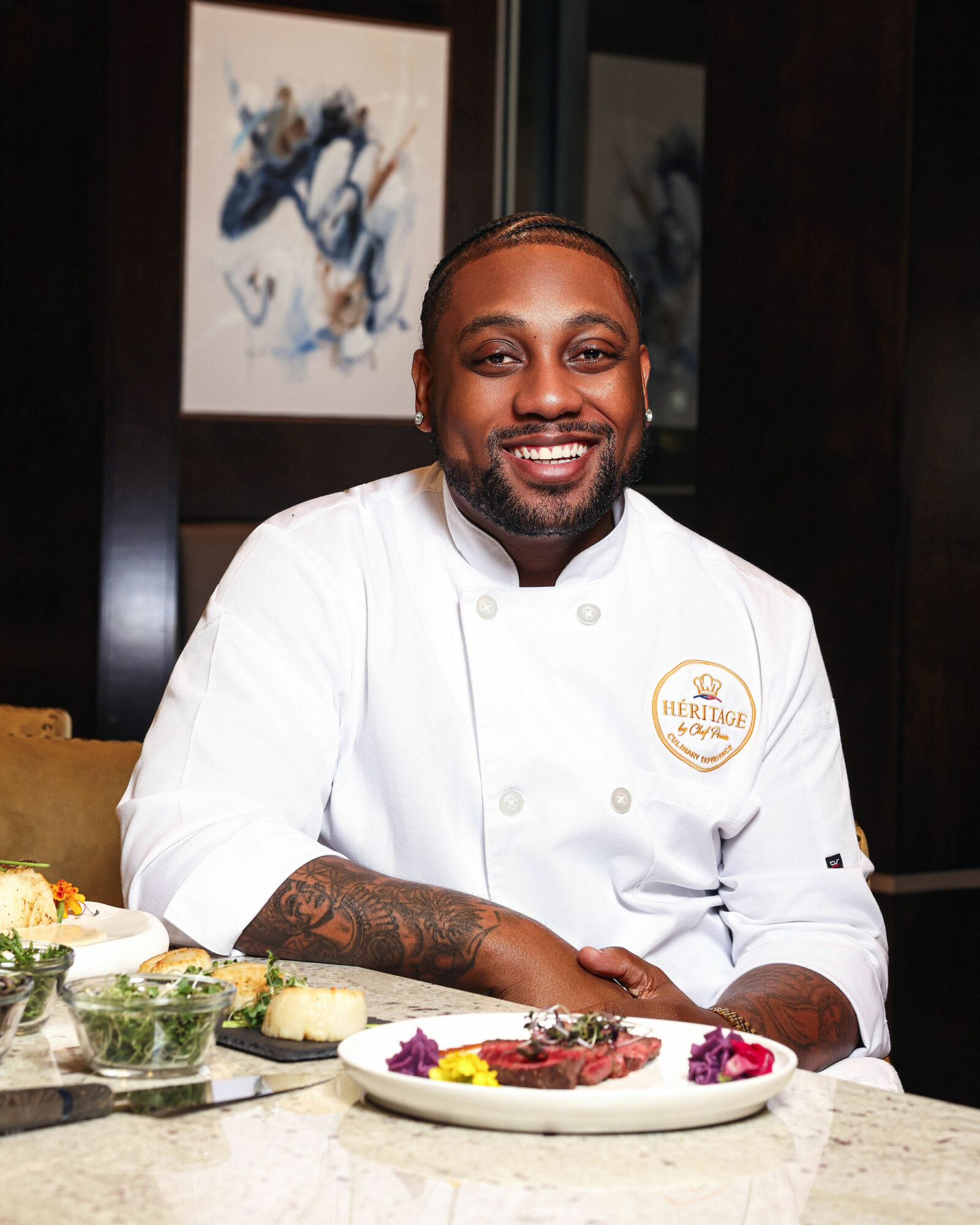 Chef in white uniform smiling, seated at a table with food, dark background with abstract art, arm tattoos visible.