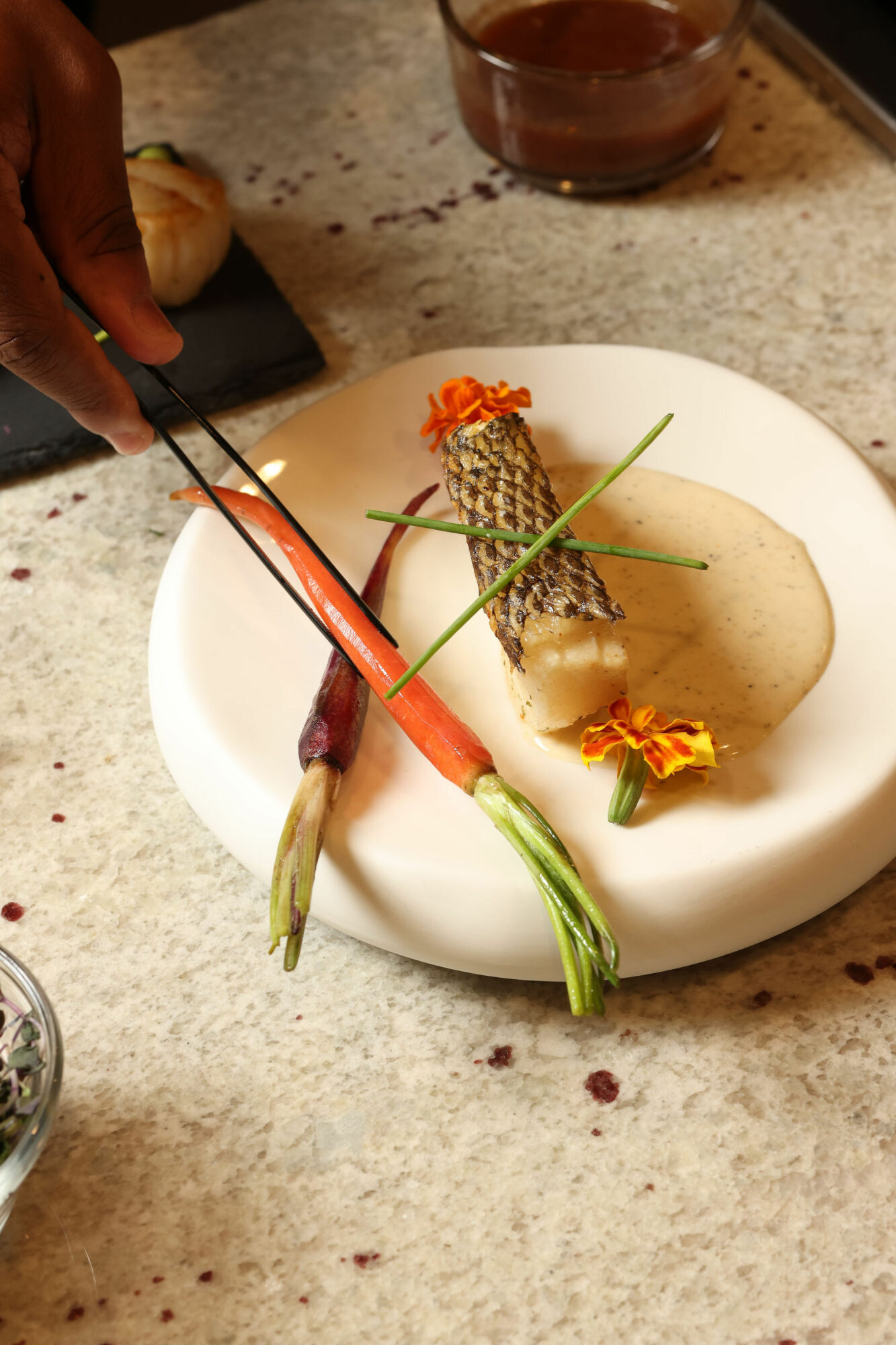 A plated dish with fish, vegetables, and garnishes on a white plate, on a textured surface, with a hand holding chopsticks.