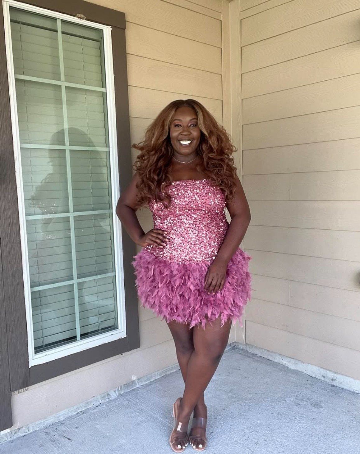 Woman in pink dress with feathered skirt standing outside near a window and beige wall, smiling, hands on hips.