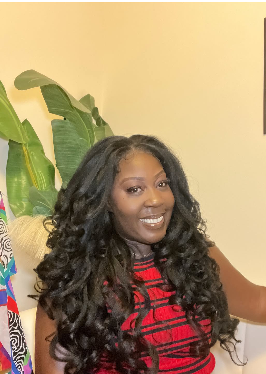 Woman with long, curly black hair smiling, wearing a red and black striped top, in front of a yellow wall and green plants.
