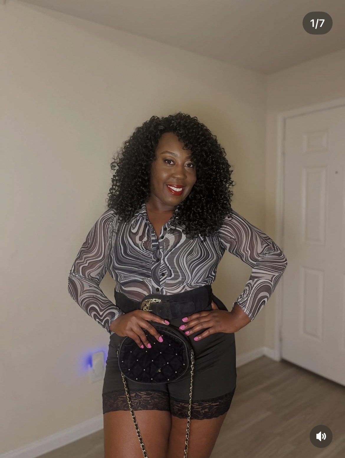 Woman with curly hair in patterned blouse and black shorts, smiling with hand on hip, standing indoors.