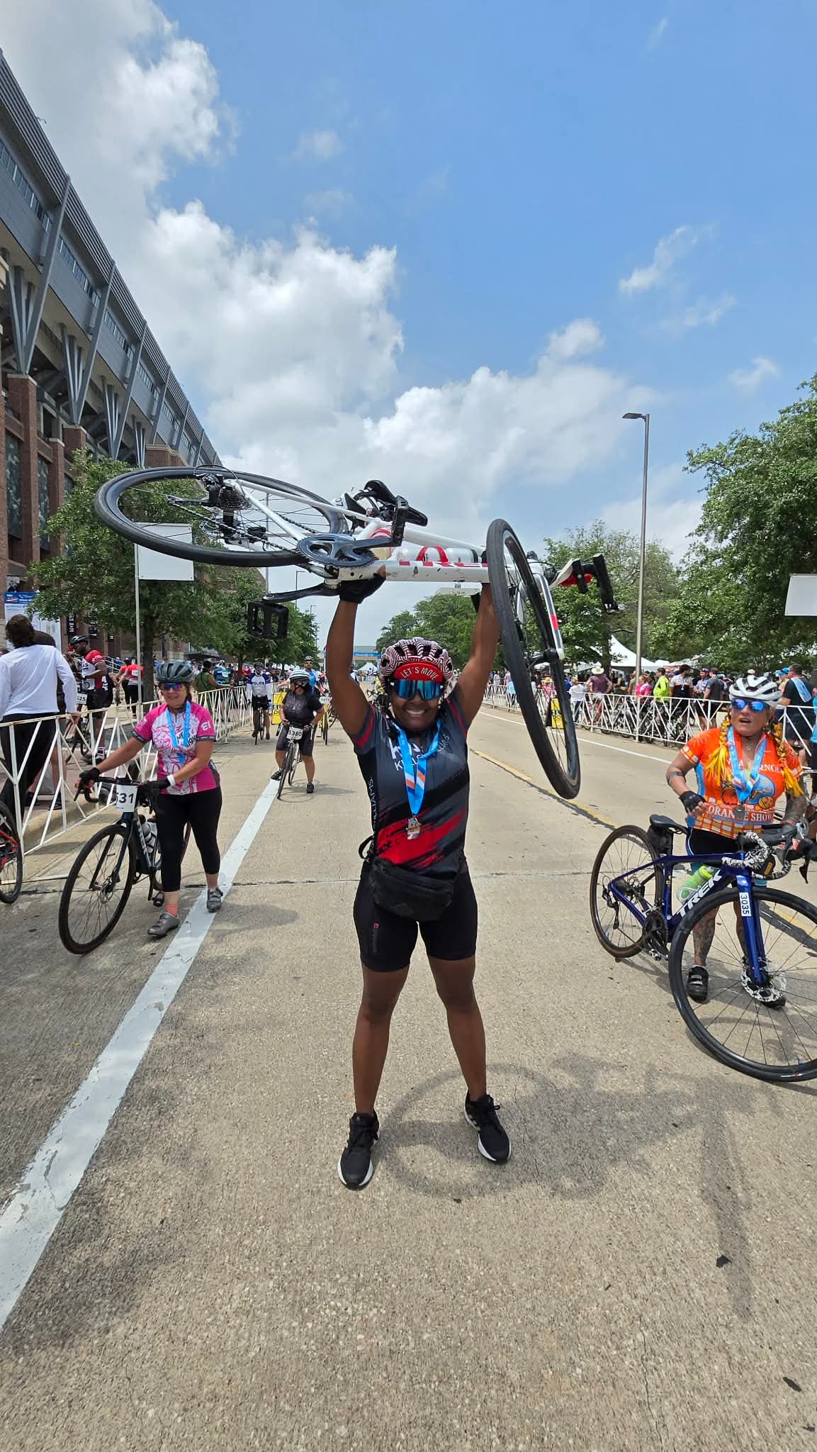 Person holding a bicycle above their head outdoors with other people and bicycles around, blue sky with clouds above.