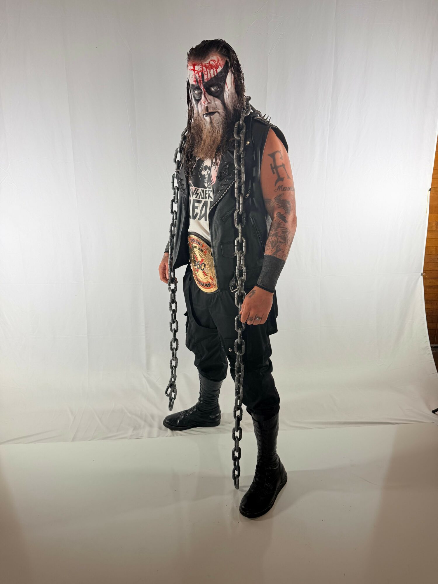 Person dressed in dark clothing with chains, makeup, and tattoos, standing against a plain white background.