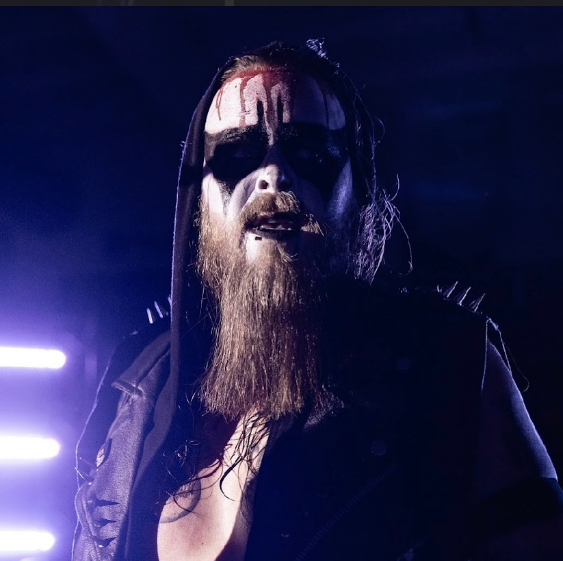 Person with black and white face paint, long hair, beard, wearing dark clothing, in a dark setting with purple lighting