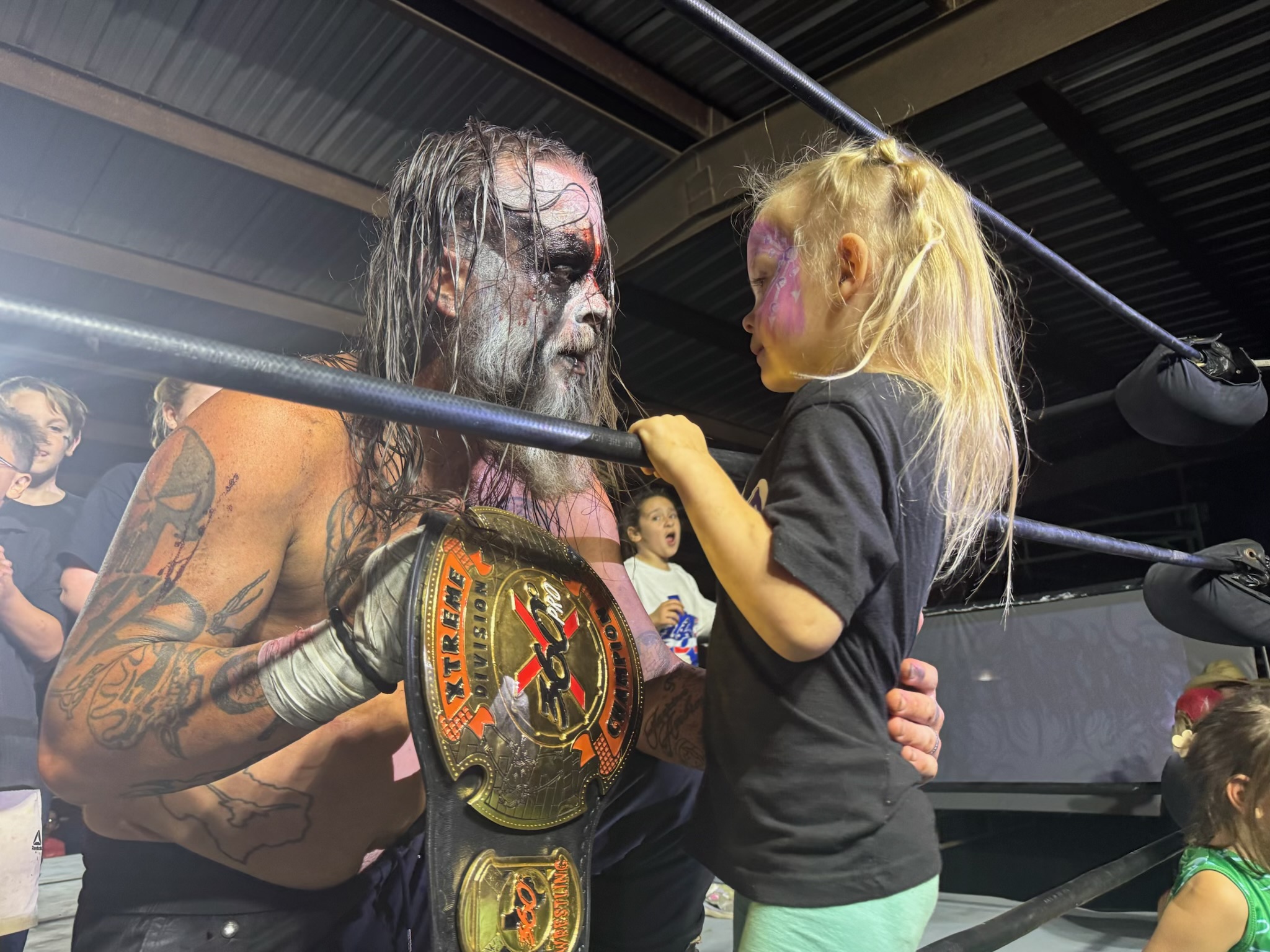 A man with face paint and tattoos holds a championship belt, facing a young girl in a black shirt inside a wrestling ring.