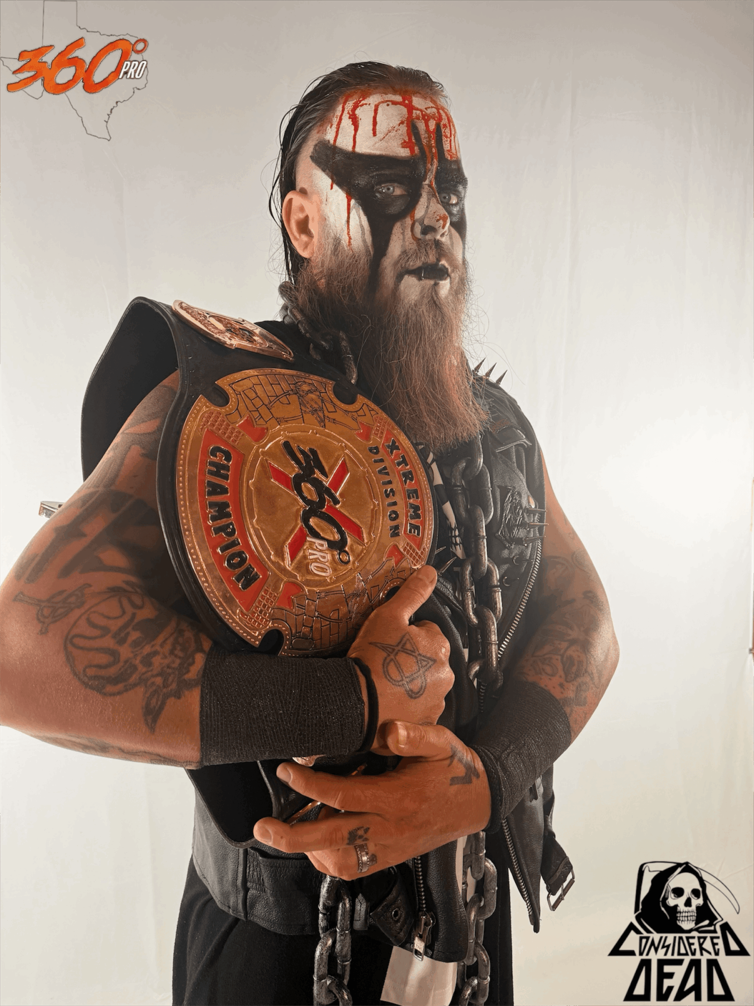Man with face paint and tattoos holding a championship belt, wearing black clothing and accessories, against a plain background.