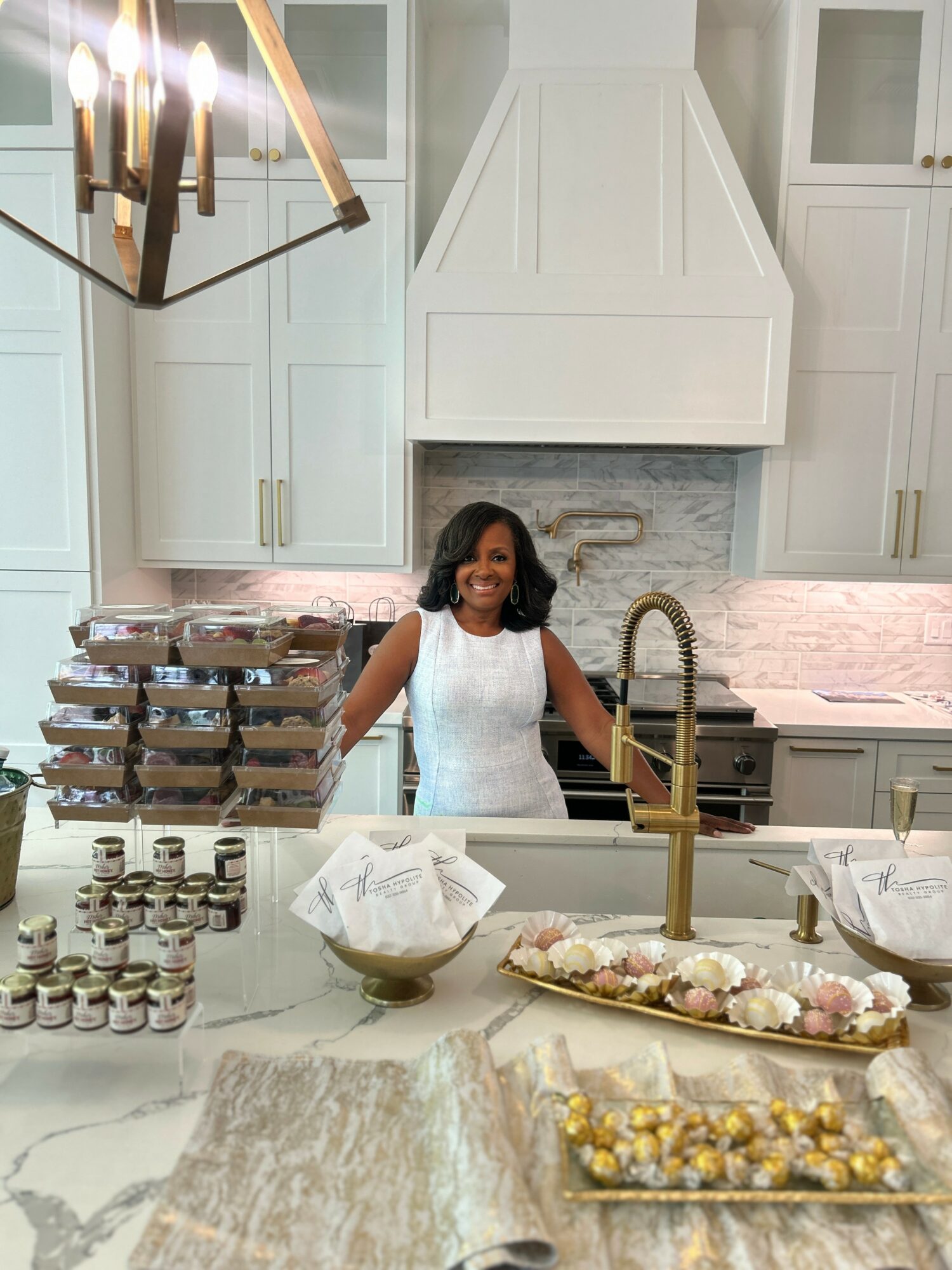 Woman standing behind kitchen island with baked goods and jars, smiling, white cabinets and range in background.