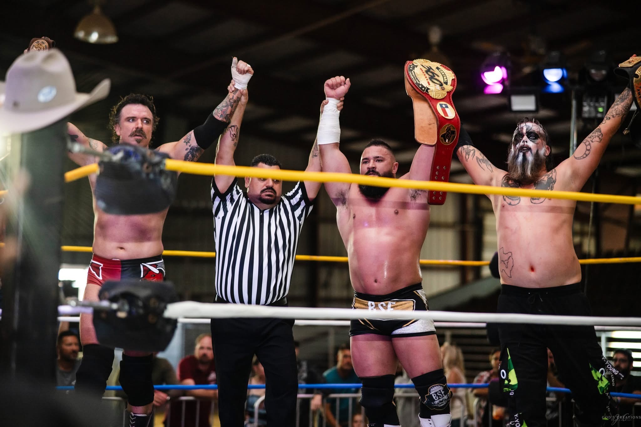 Three wrestlers and a referee in a wrestling ring, one wrestler holding a championship belt, celebrating victory.