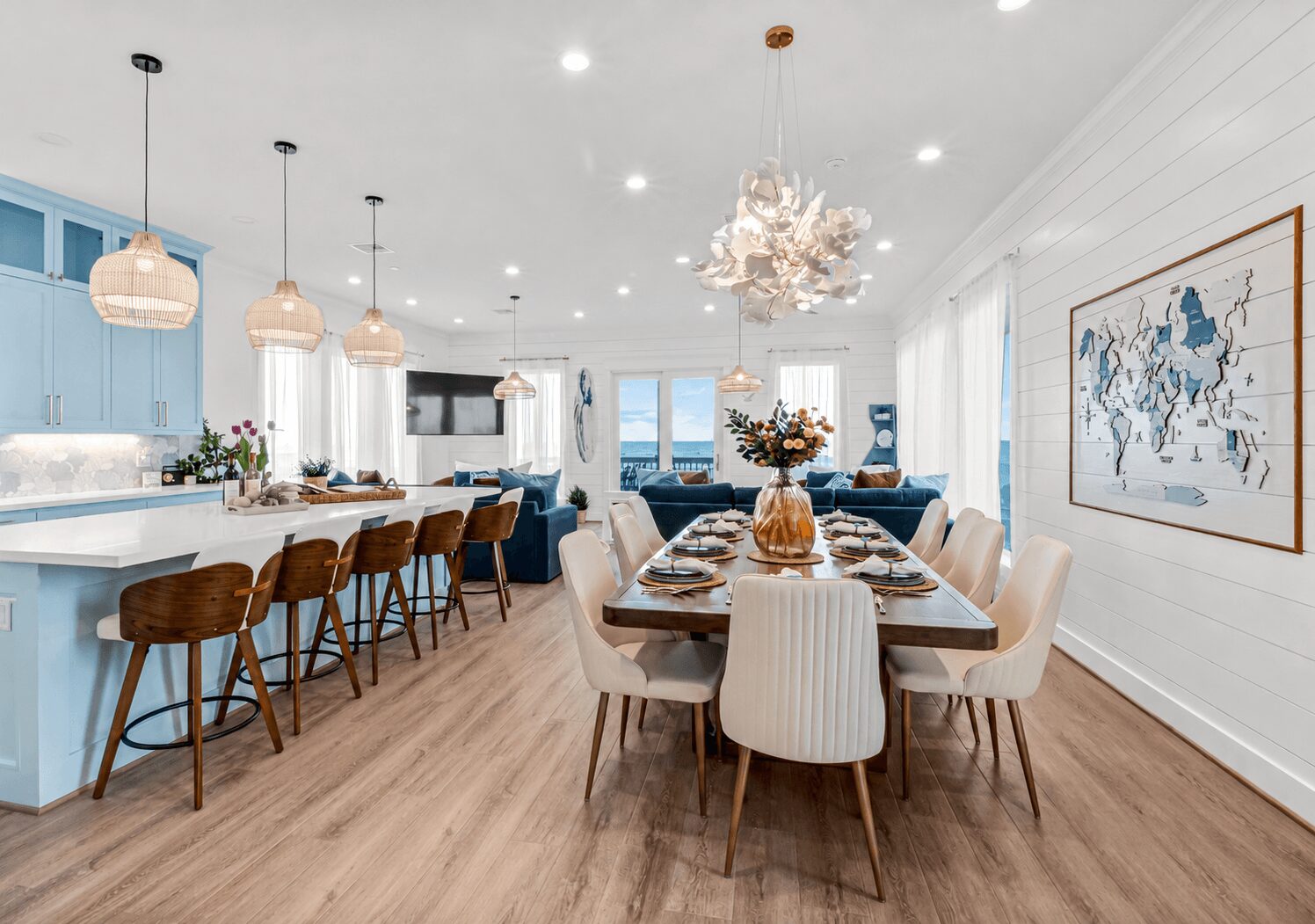 Open-concept dining and kitchen area with a long table, beige chairs, blue kitchen cabinets, and pendant lights, bright and airy.