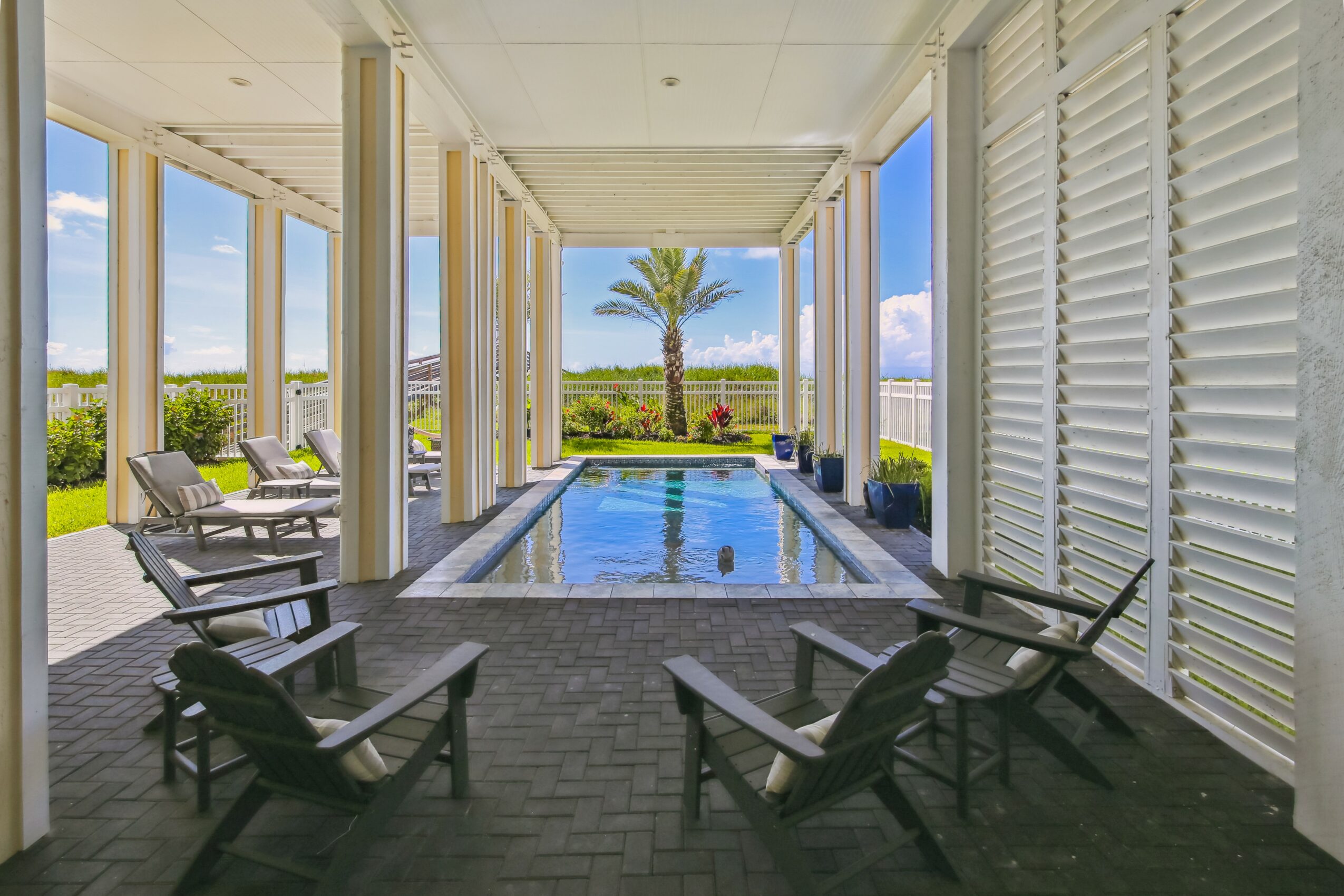 Indoor pool area with chairs, large windows, and a view of a palm tree outside, bright and sunny.