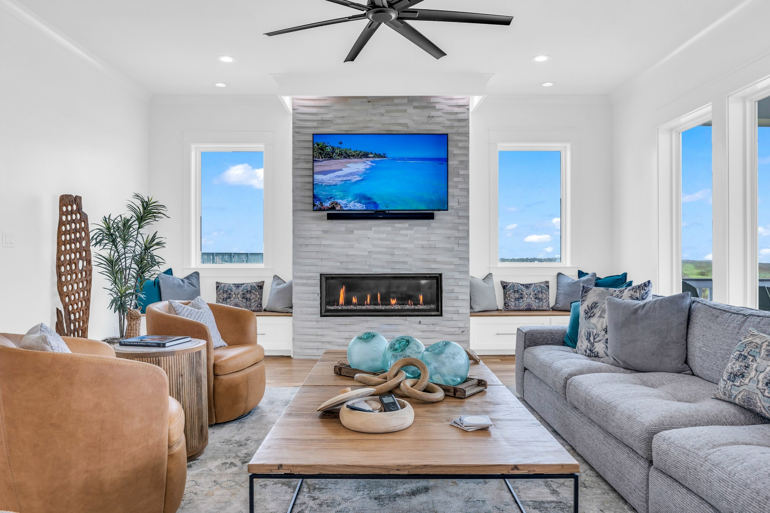 Living room with a fireplace, wall-mounted TV, large windows, and various sofas and chairs, decorated with plants and cushions.