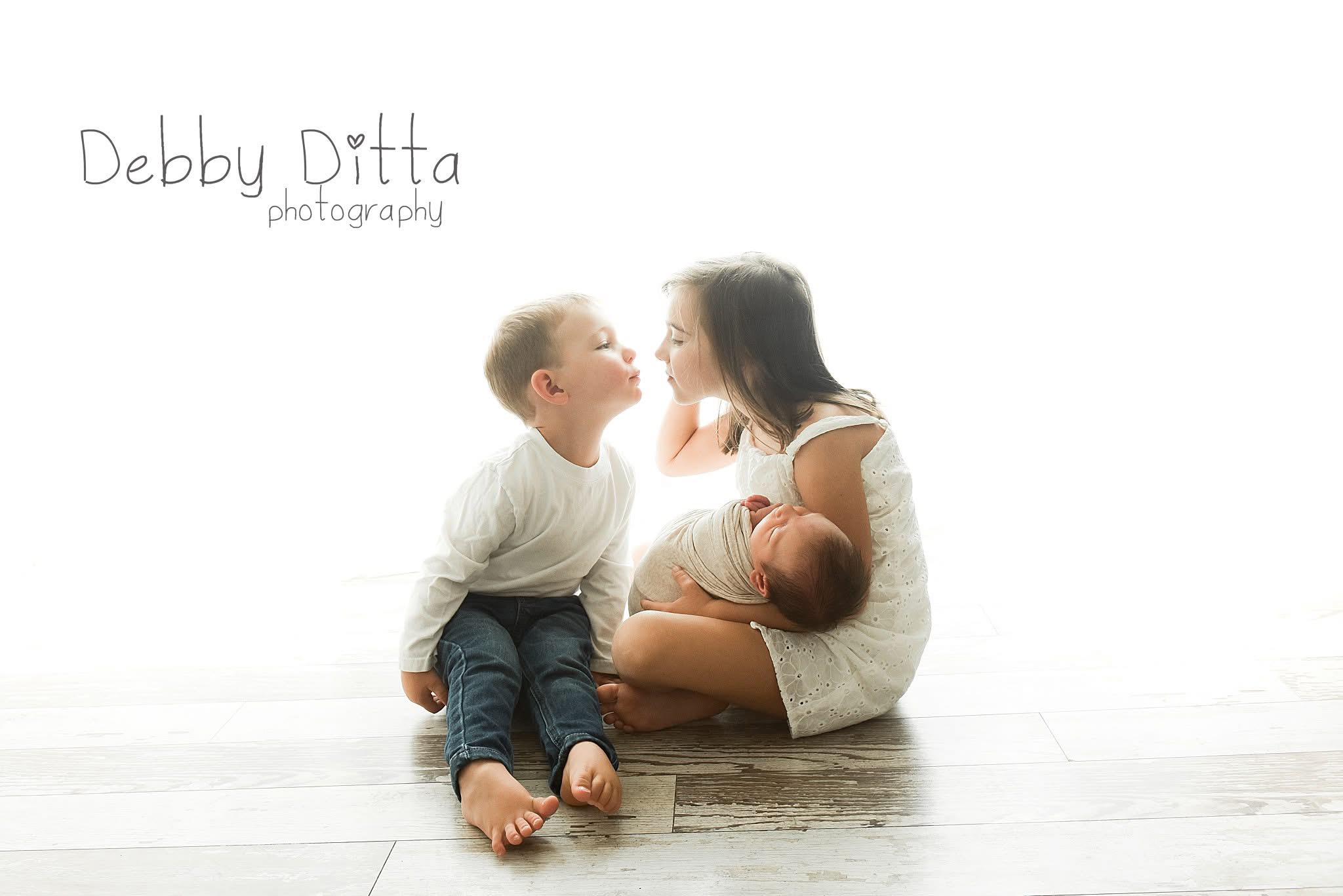 A woman with a baby and a young boy sitting on a wooden floor, facing each other, with a white background.
