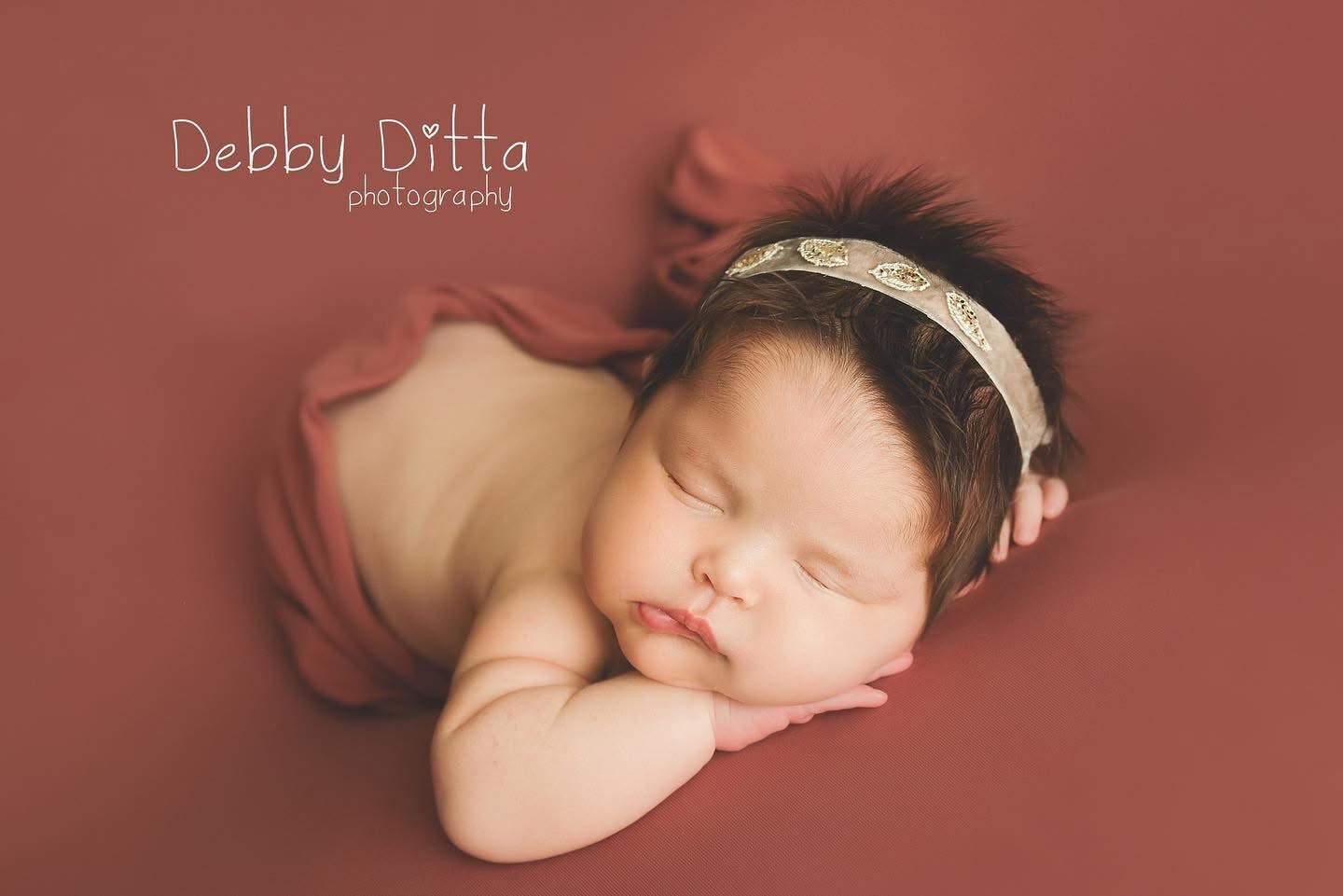 Young baby with a headband, sleeping on a soft surface, resting face on hand, with a pink background.