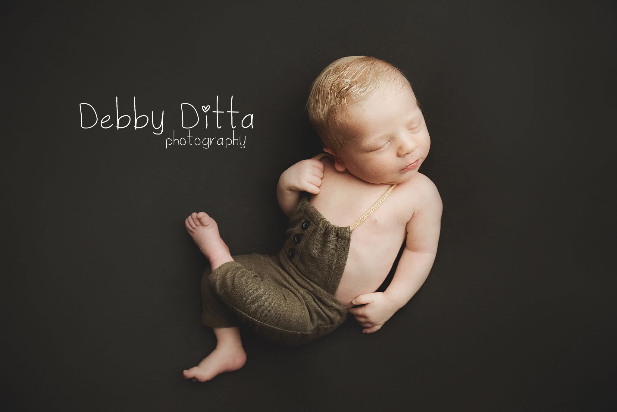 Baby lying on dark background, wearing olive-green pants, with head turned to the side, eyes closed, and arms bent.