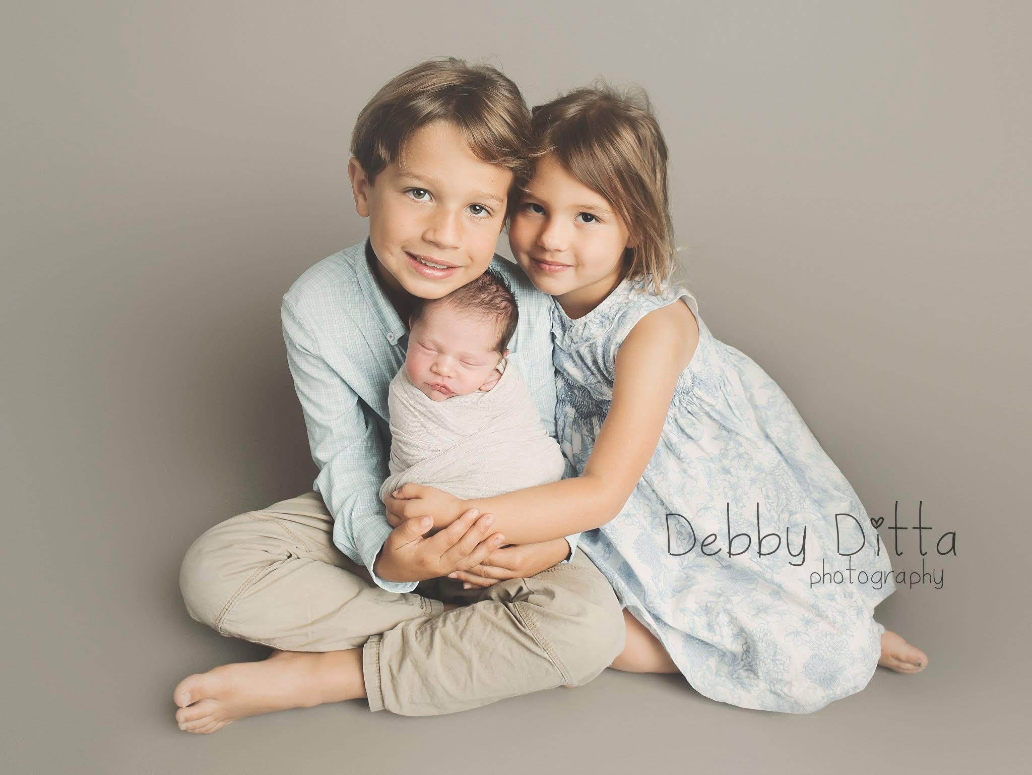 Two children, a boy and a girl, sit with a newborn baby on a plain background, smiling and hugging.