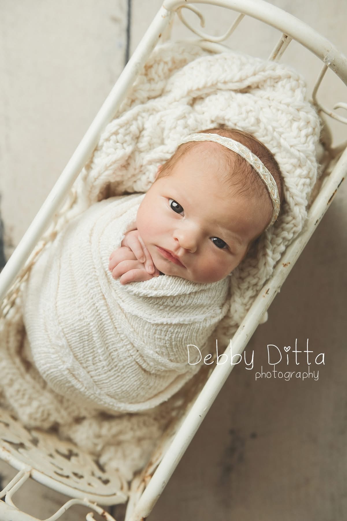 Baby lying in a crib, wrapped in a white blanket, with a headband and a fluffy blanket behind.
