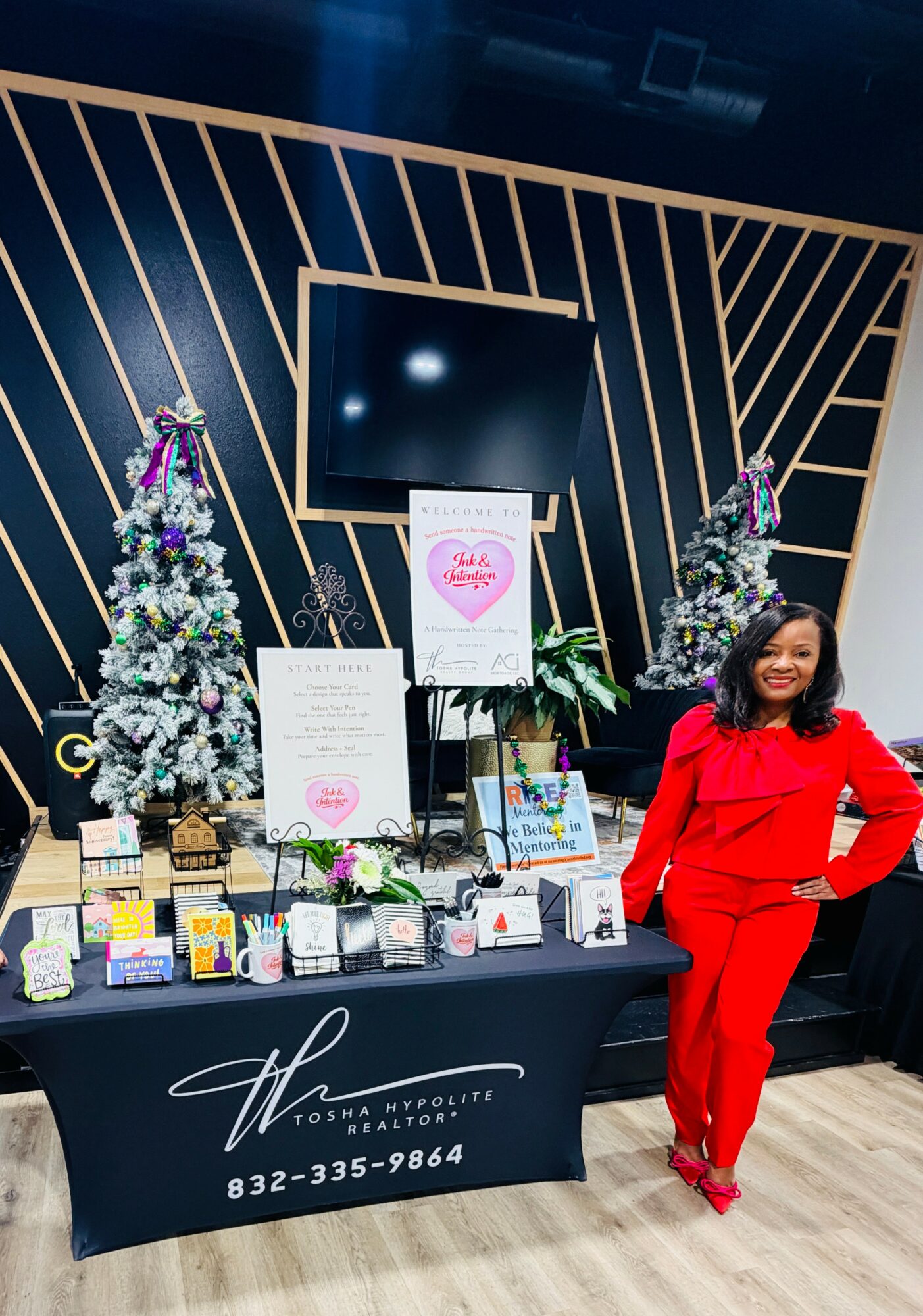 Woman in red outfit standing next to a table with holiday decorations and products, with a decorated Christmas tree in the background.