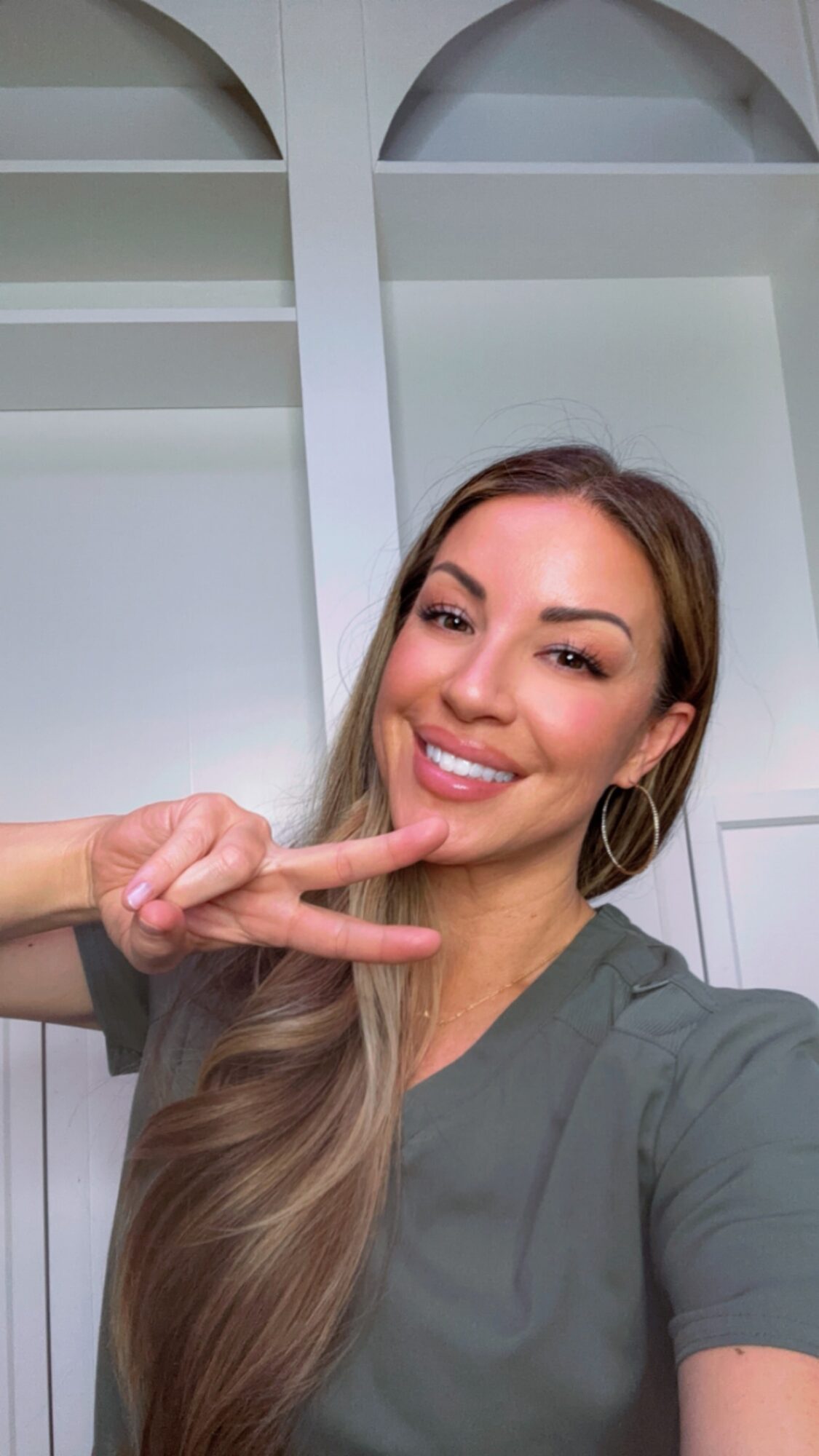 Young woman with long hair smiling and making a peace sign gesture, indoors with white shelves in background.