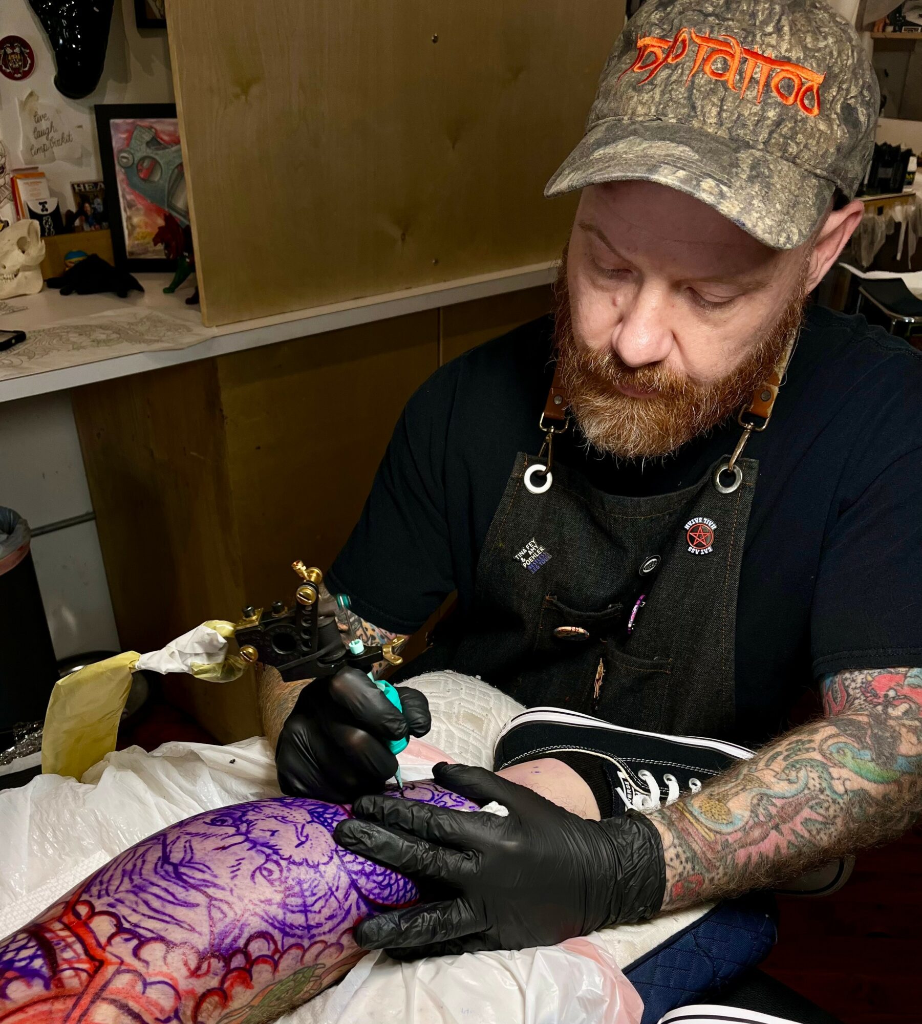 Tattoo artist with a beard, wearing a camouflage cap, working on a purple tattoo on a person's arm in a studio.