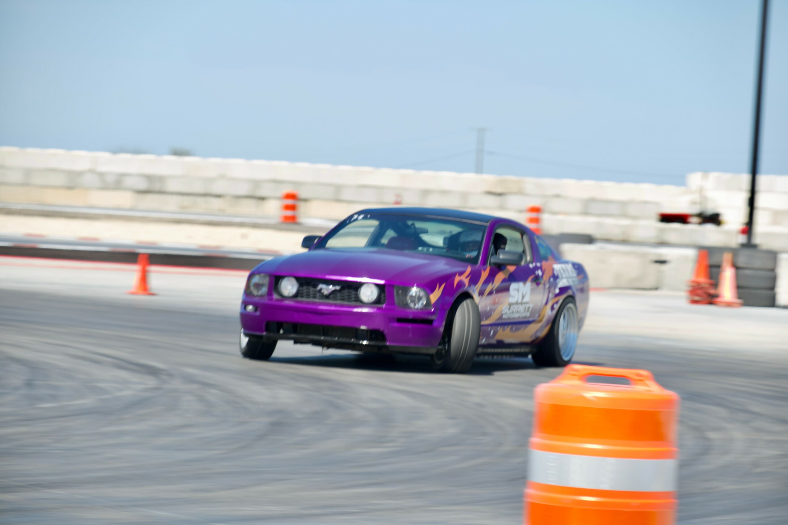 Purple sports car drifting on a track with orange cones, concrete barriers, and a clear blue sky.