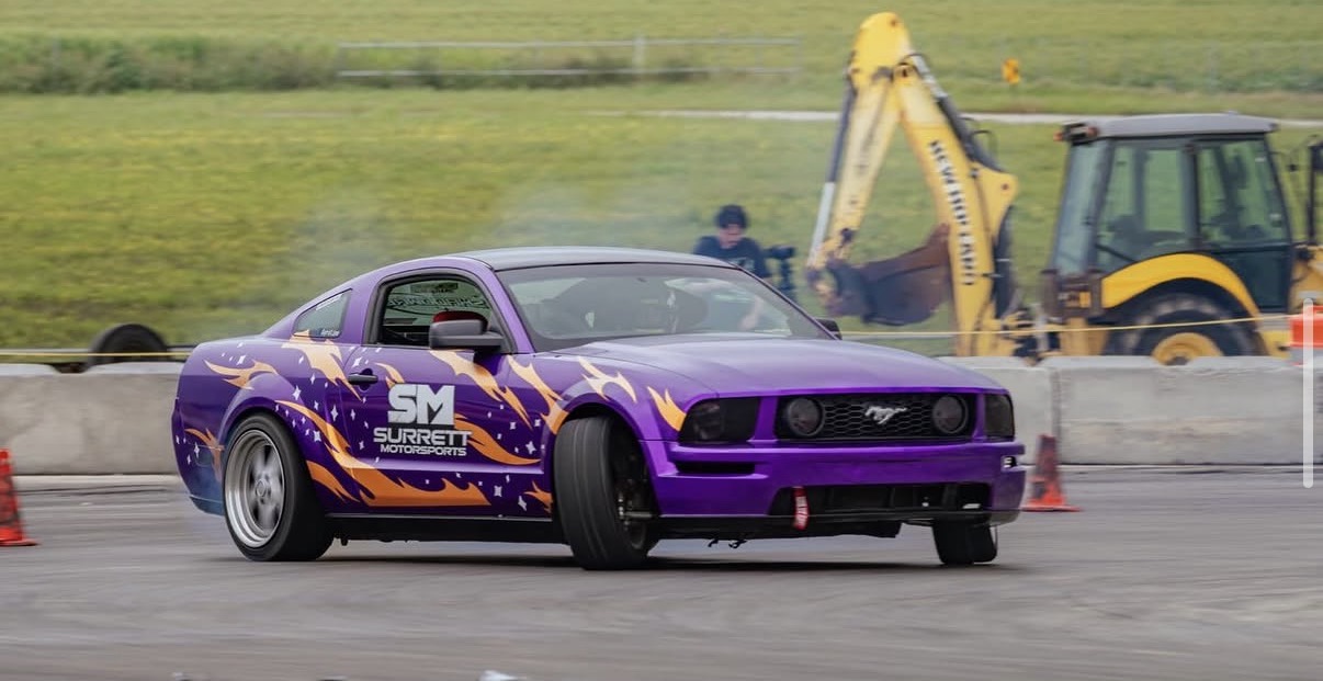 Purple race car with orange and yellow graphics on a track, construction equipment in background, cones on the ground.