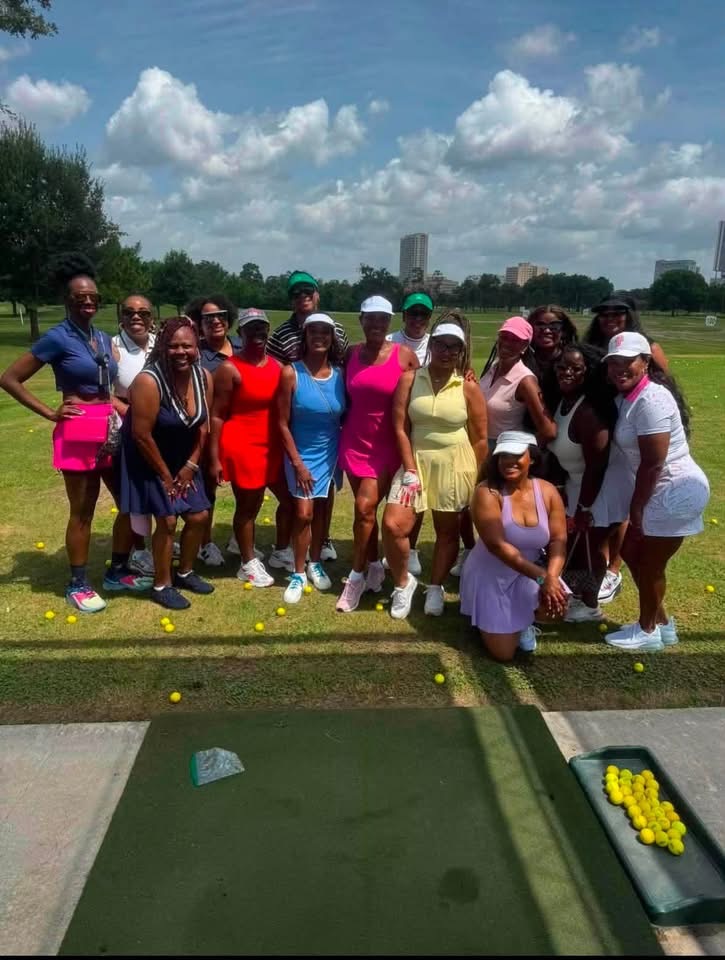 Group of women and children at a golf course, standing on grass with golf balls and a tray of yellow balls, under a partly cloudy sky.