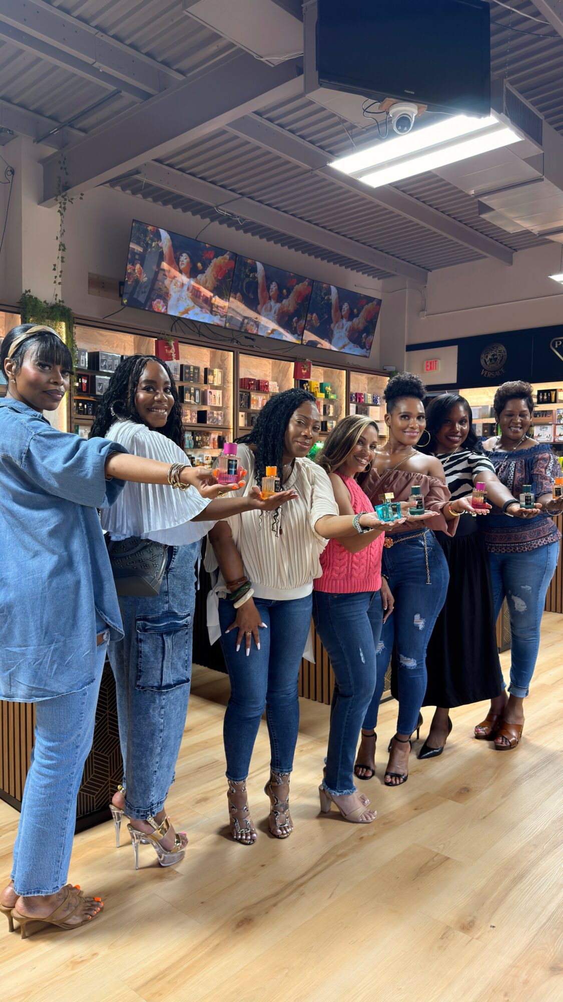 Group of women standing in a line inside a store, holding drinks and smiling, with shelves and a TV in the background.
