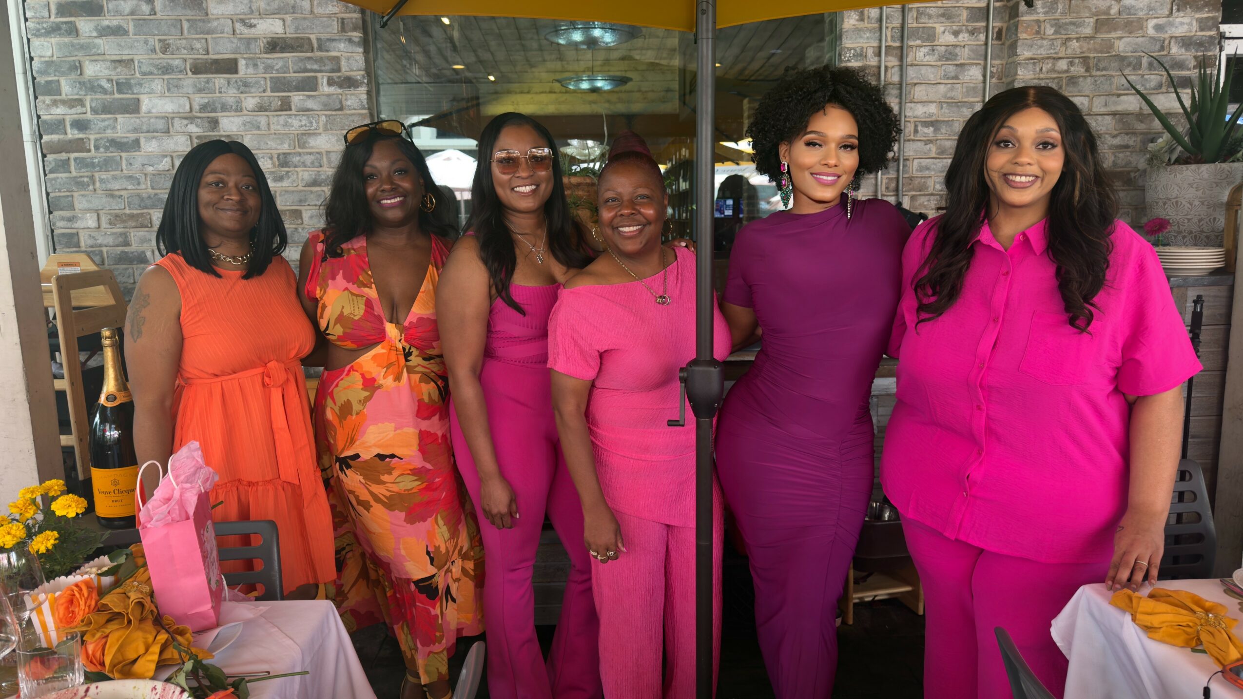 Six women standing together indoors, smiling, wearing colorful dresses and outfits, with tables and decorations nearby.