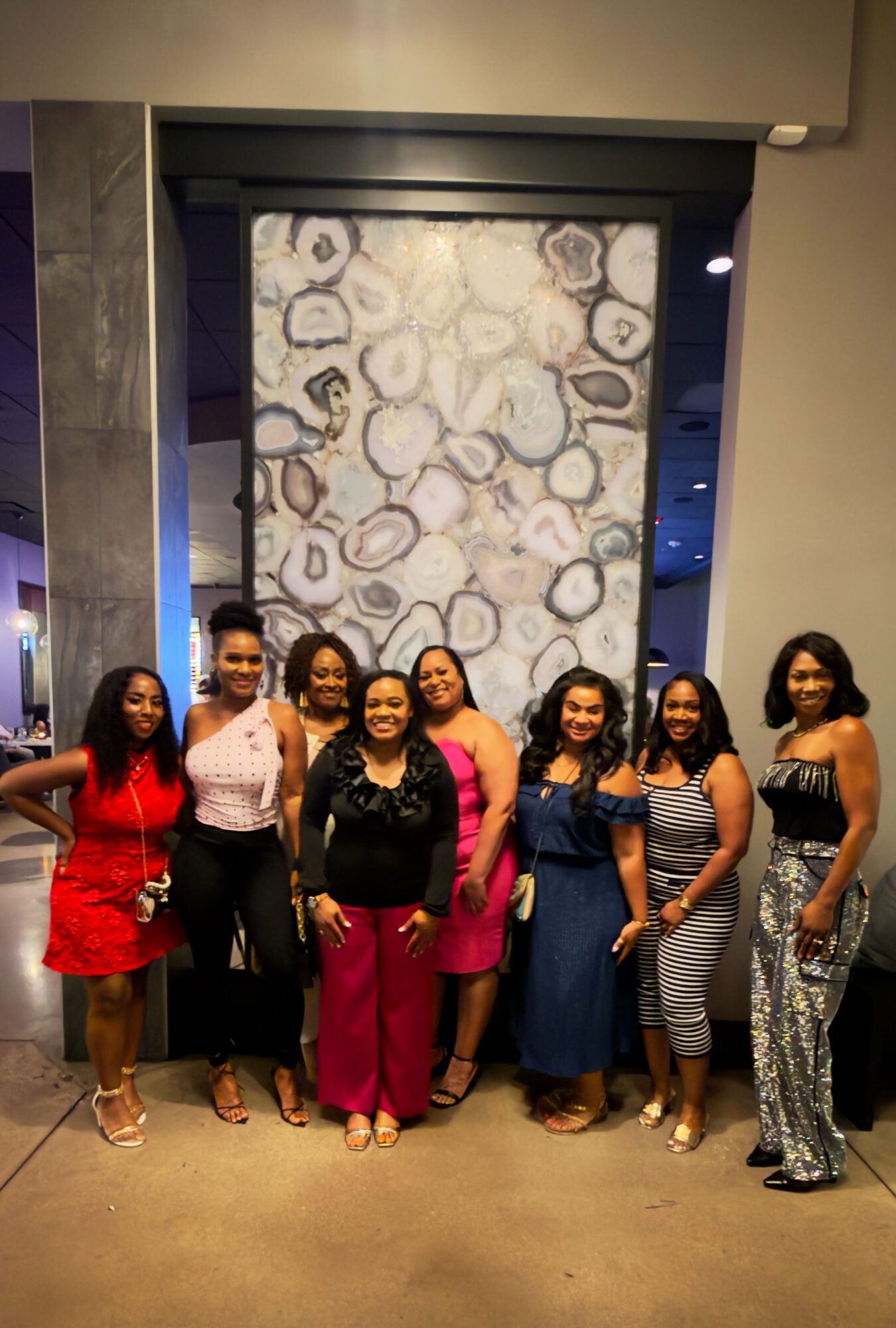 Group of nine women standing together indoors in front of a decorative wall, smiling, dressed in colorful outfits.