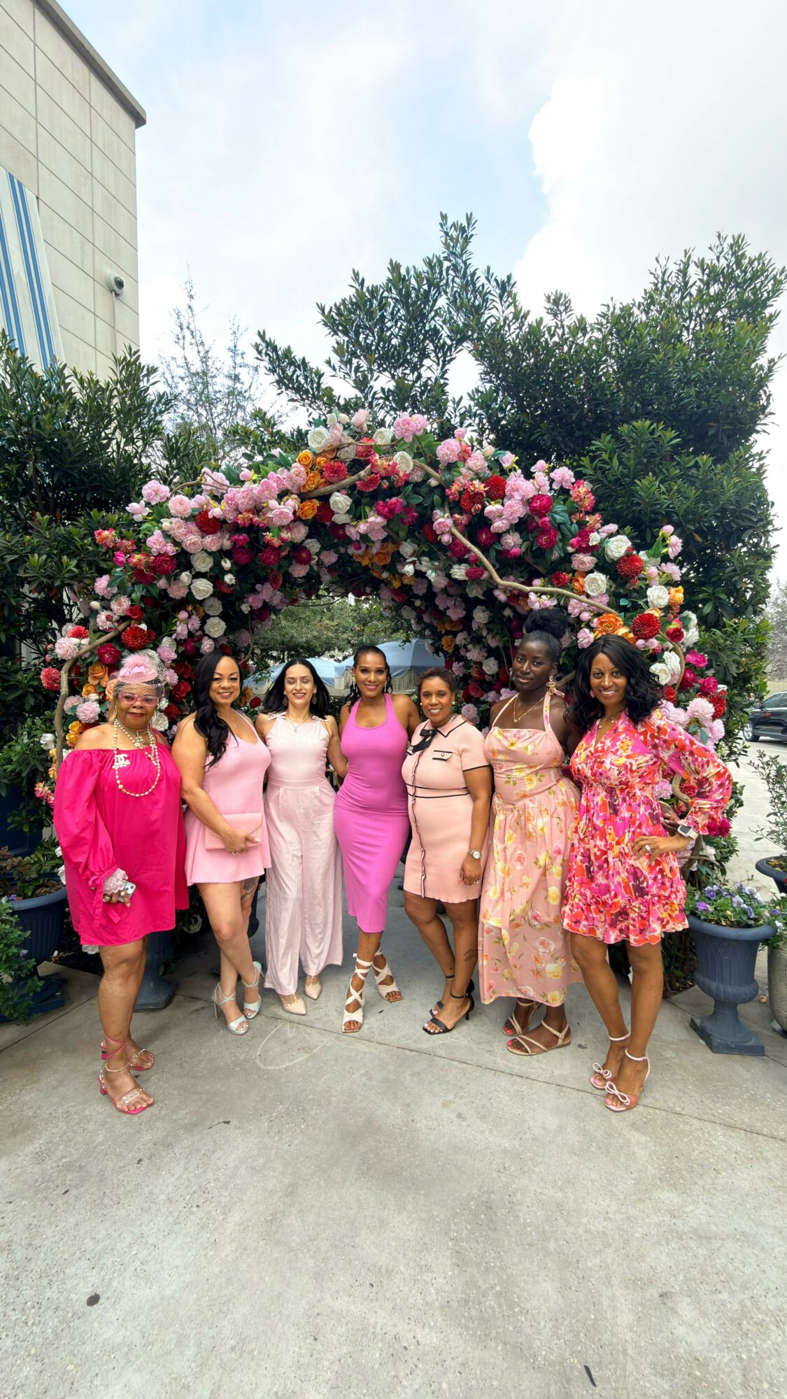 Six women stand under a floral arch with pink, white, and red flowers outdoors, with trees and a building in the background.